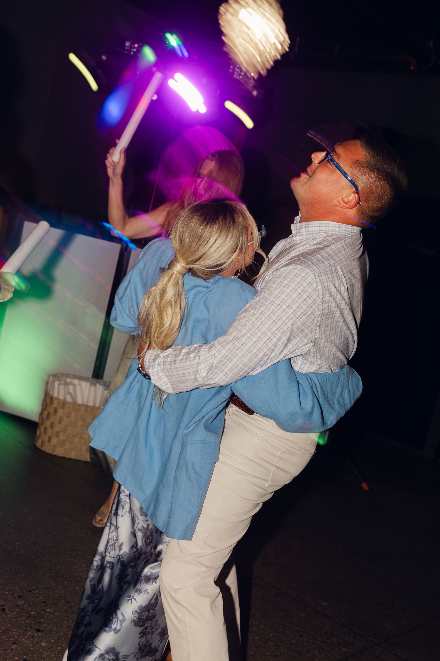 Guests dance and embrace under colorful party lights with glow sticks at a lively wedding reception.