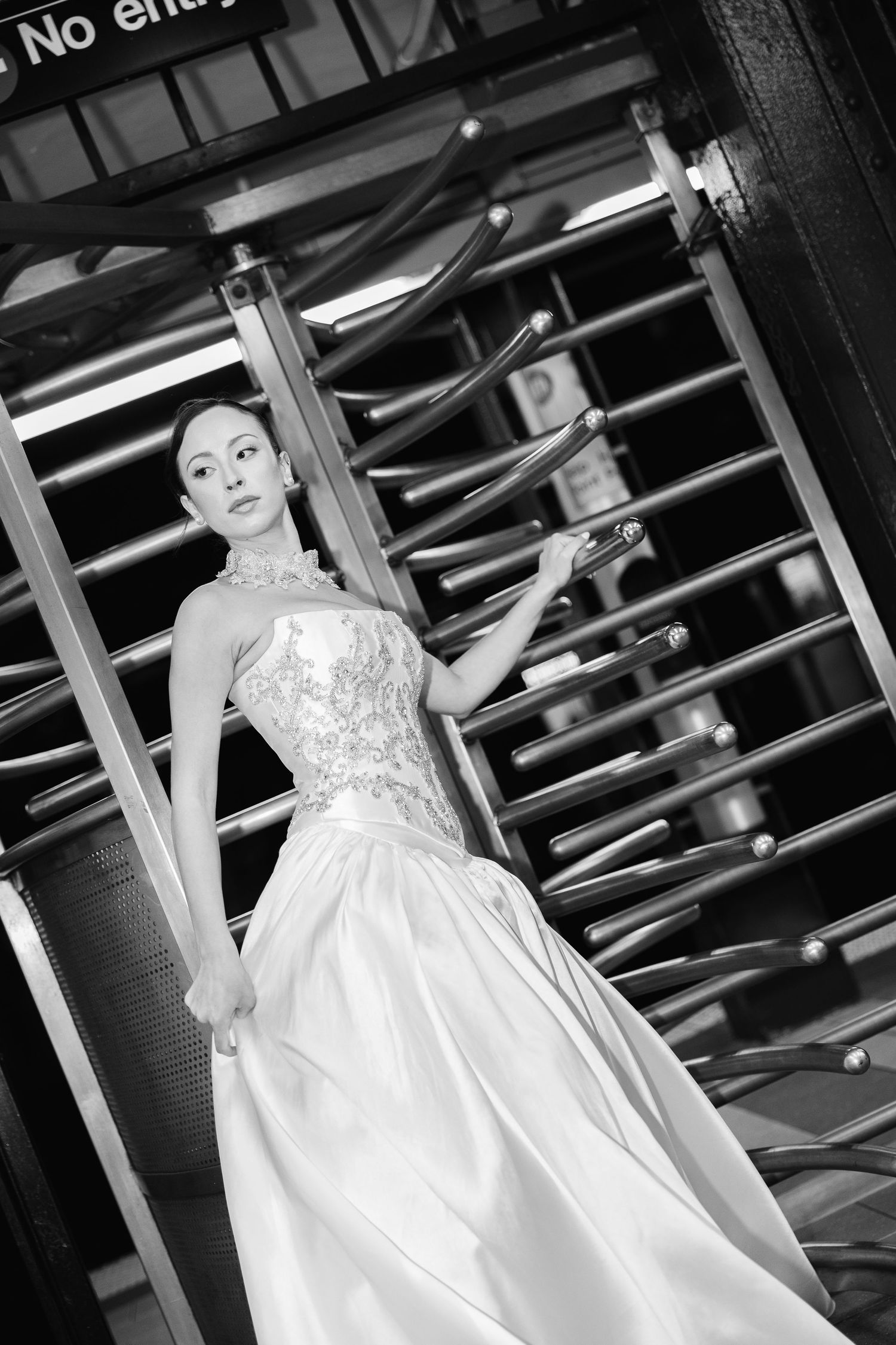 Black and white photo of a bride in a beaded strapless wedding gown posing beside a subway turnstile.