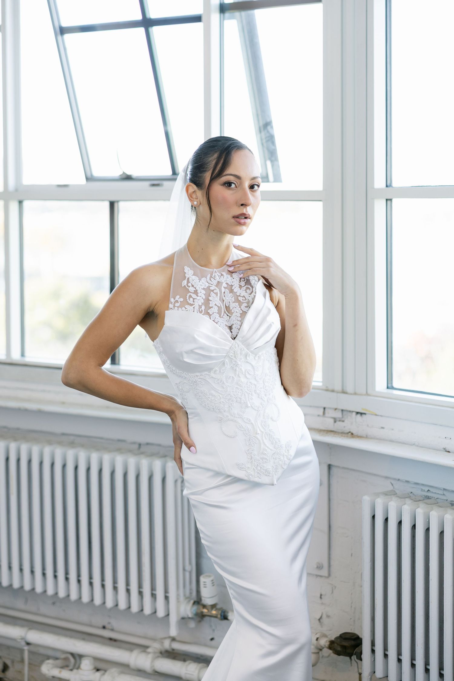 Bride in a lace halter satin wedding gown poses with her hand on her hip in front of large industrial windows.