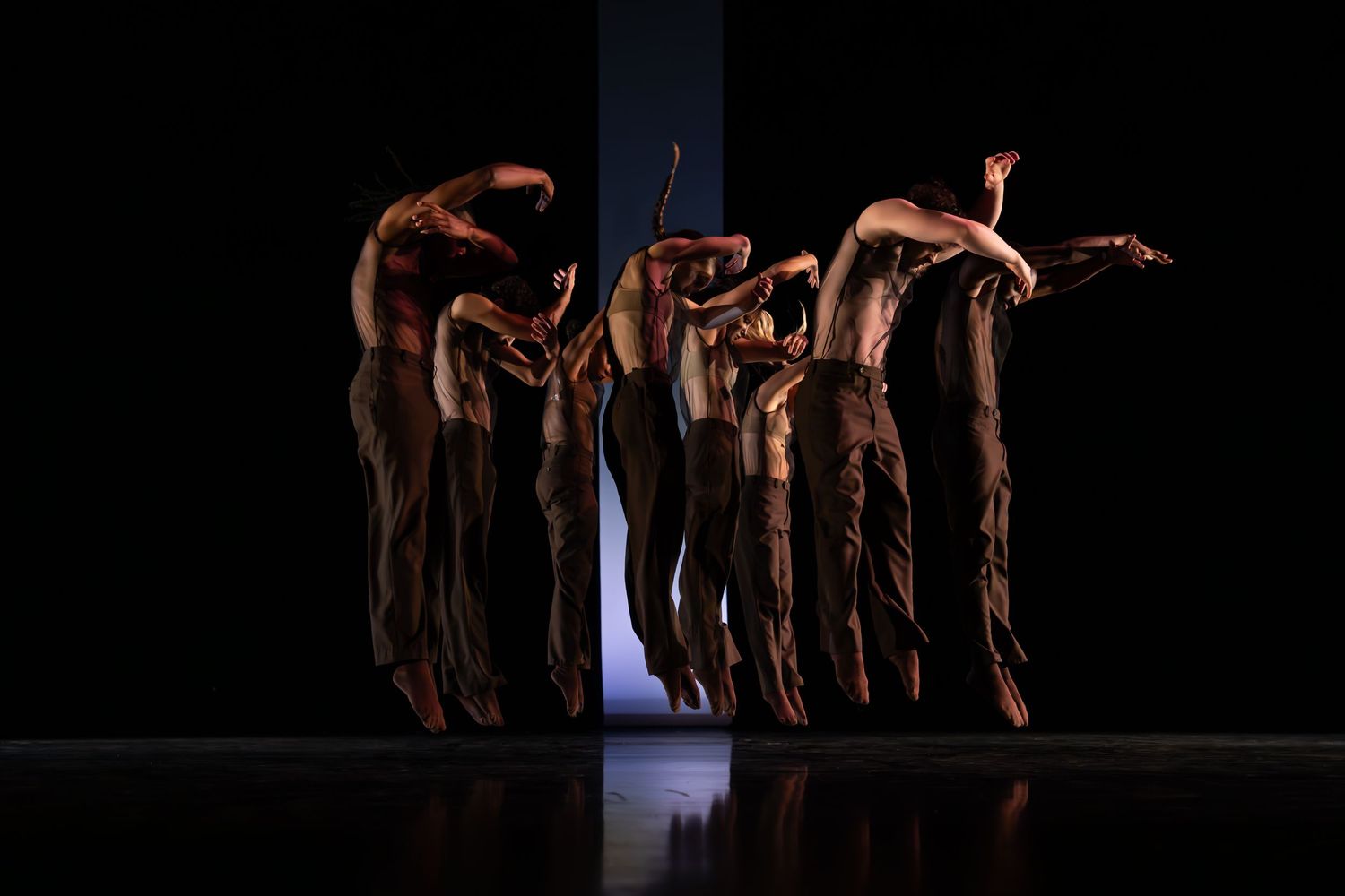 A group of dancers in loose pants stand close together on stage, bending and reaching in a striking silhouette against a dark backdrop.