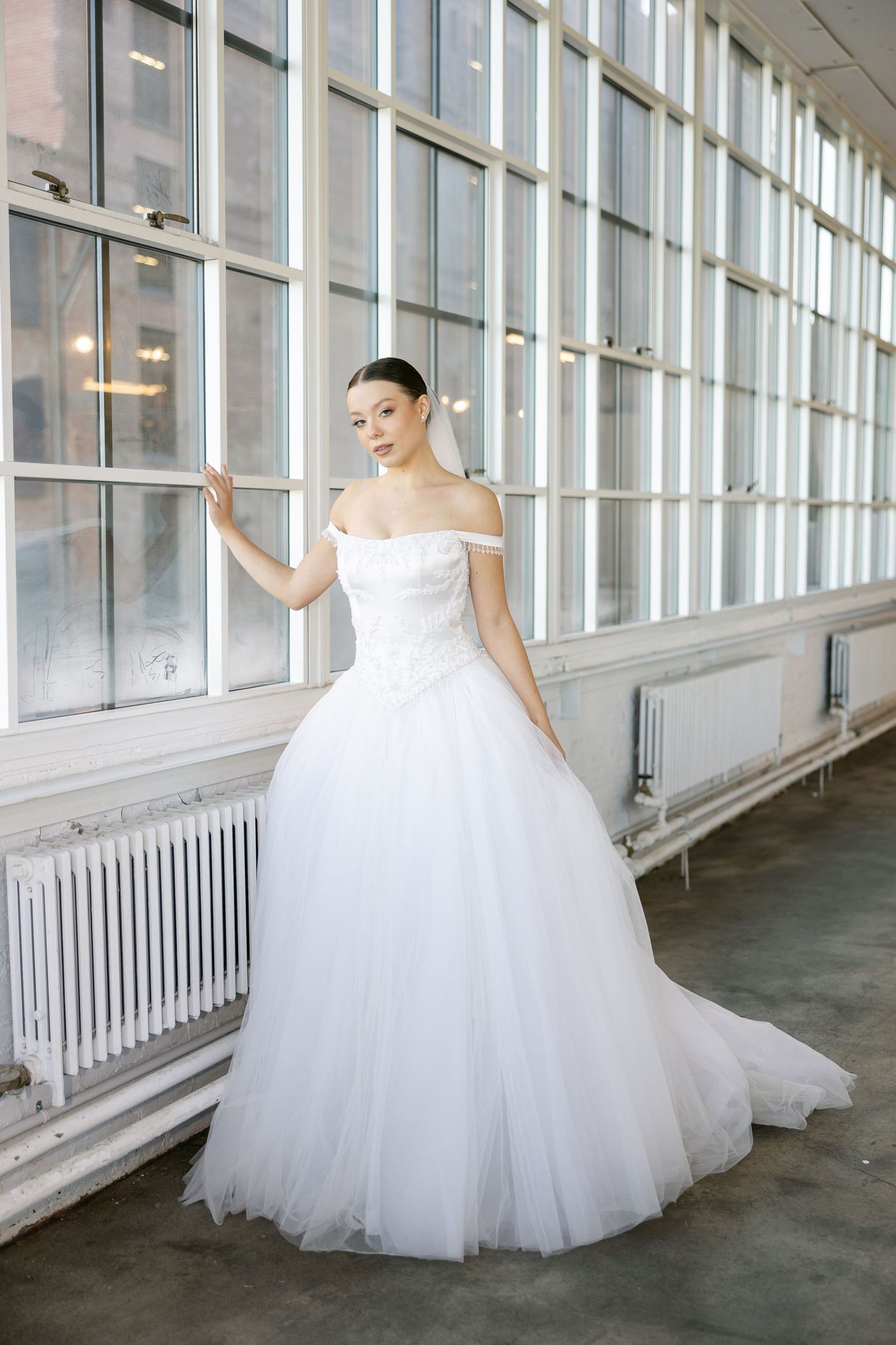 Bride in an off-the-shoulder white tulle ball gown poses beside tall industrial windows and white radiators.