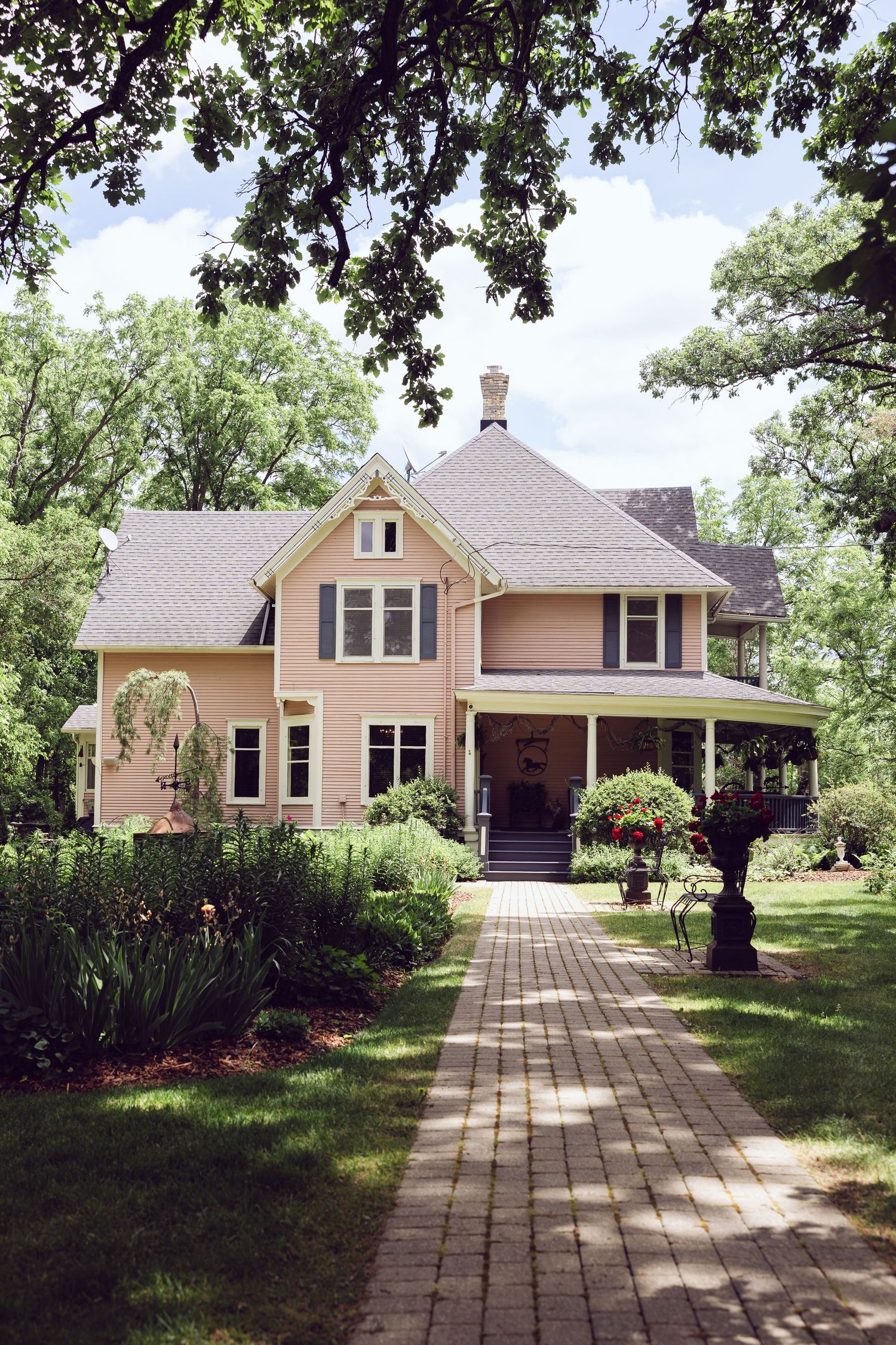 Pink Victorian farmhouse with a brick walkway, lush gardens and mature trees, a charming outdoor wedding venue in summer.