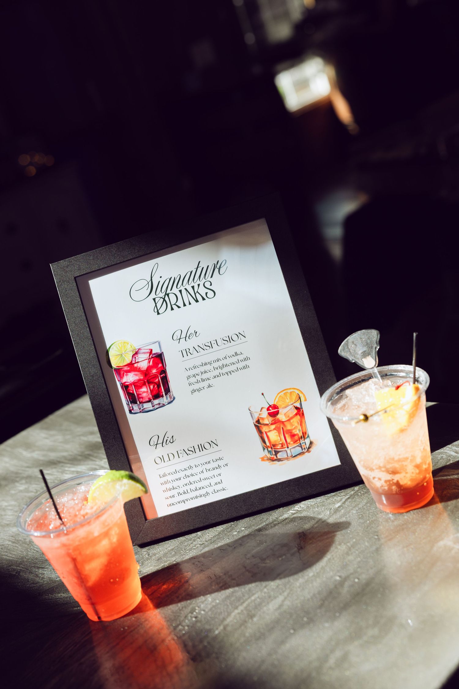Signature drinks sign listing his and her cocktails sits on a bar beside two colorful iced drinks in plastic cups.