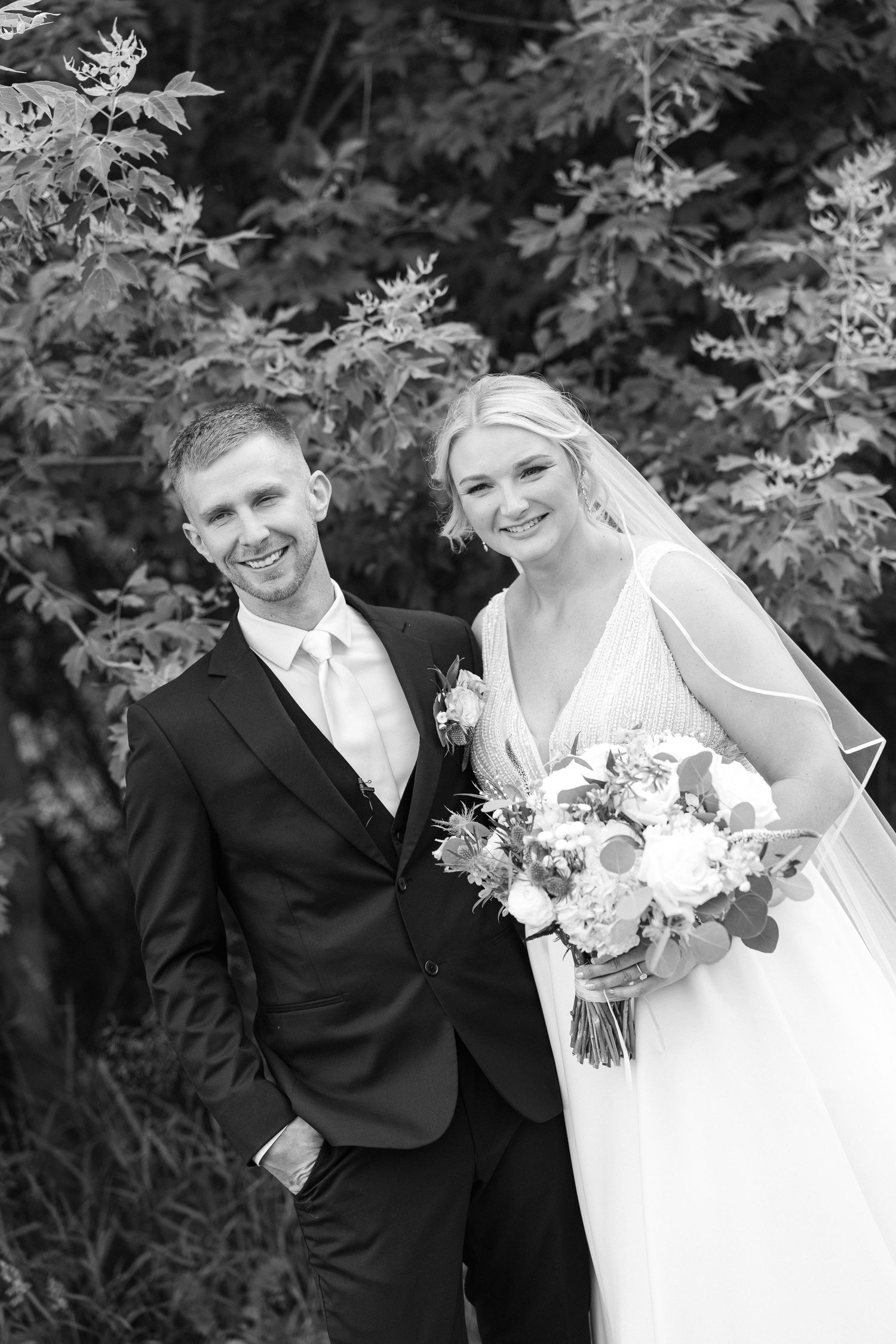 Black and white portrait of a smiling bride and groom holding a bouquet, posed among leafy trees at their wedding.