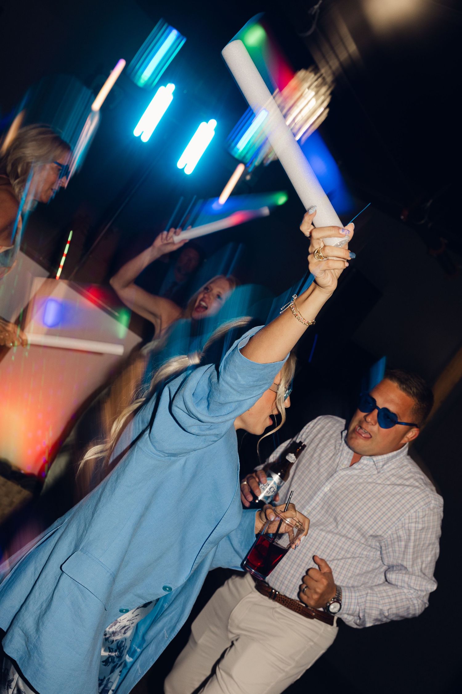 Guests dance at a wedding reception with glowing light sticks, blue and orange party lights, and drinks in hand.
