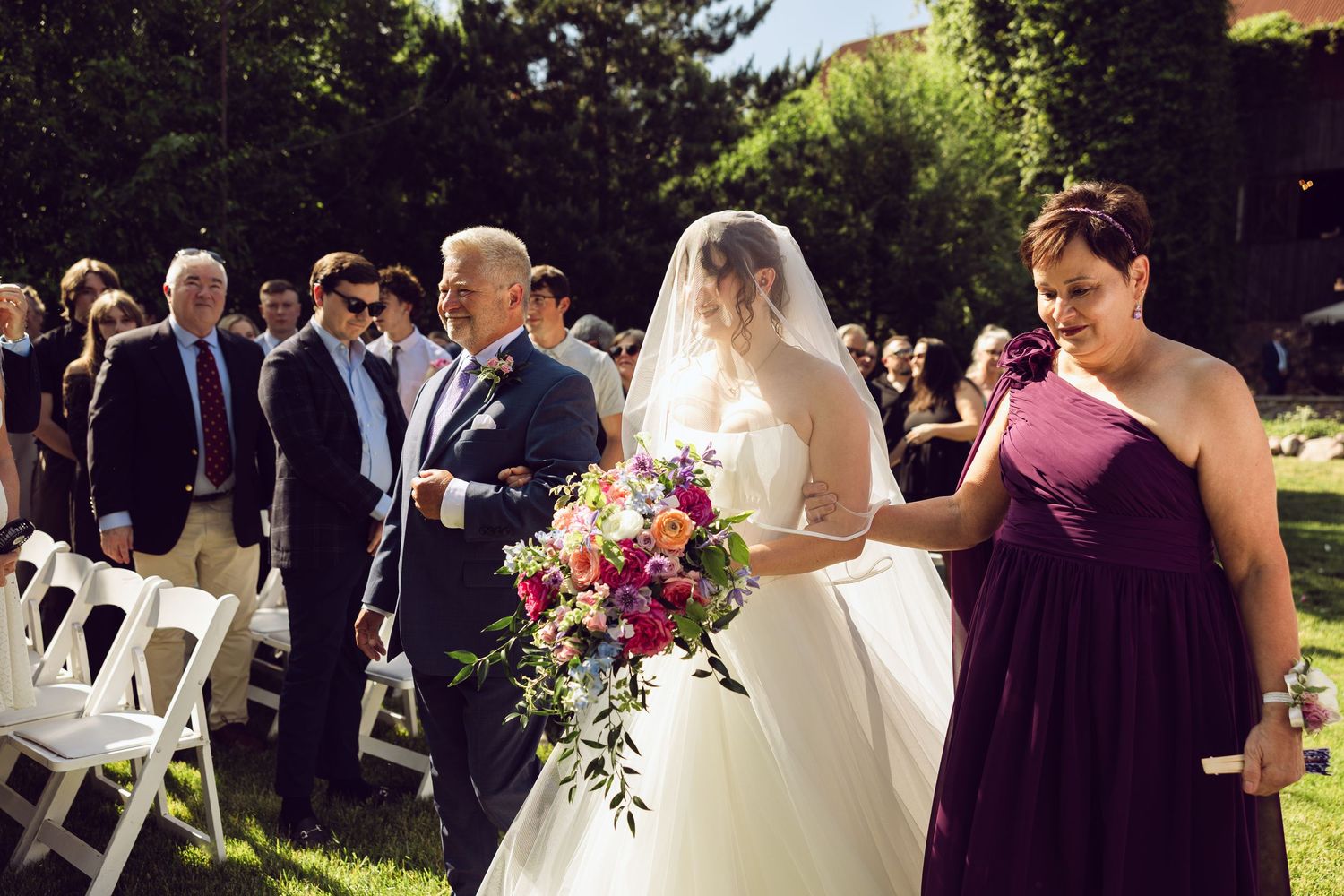 Bride in a white gown carrying a bright bouquet walks down the aisle with an older man as guests watch at an outdoor ceremony.