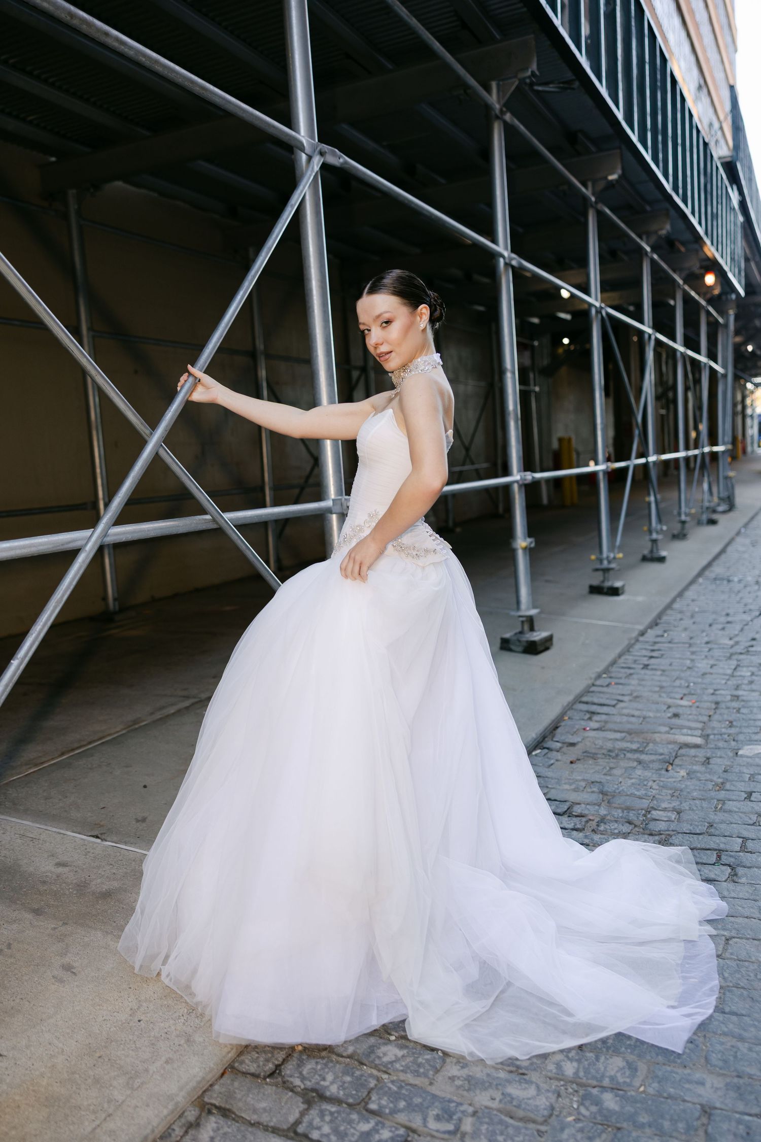 Bride in a flowing tulle wedding gown with a long train stands beside street scaffolding on a cobblestone road.