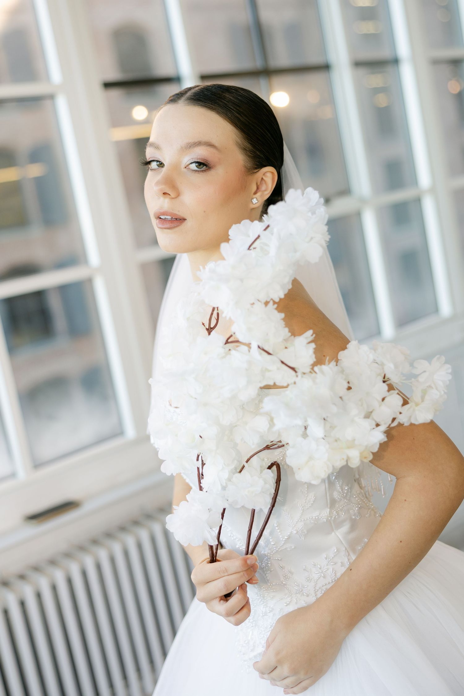 Bride in a lace bodice wedding gown and sheer veil holds a bouquet of white blossom branches by a window.