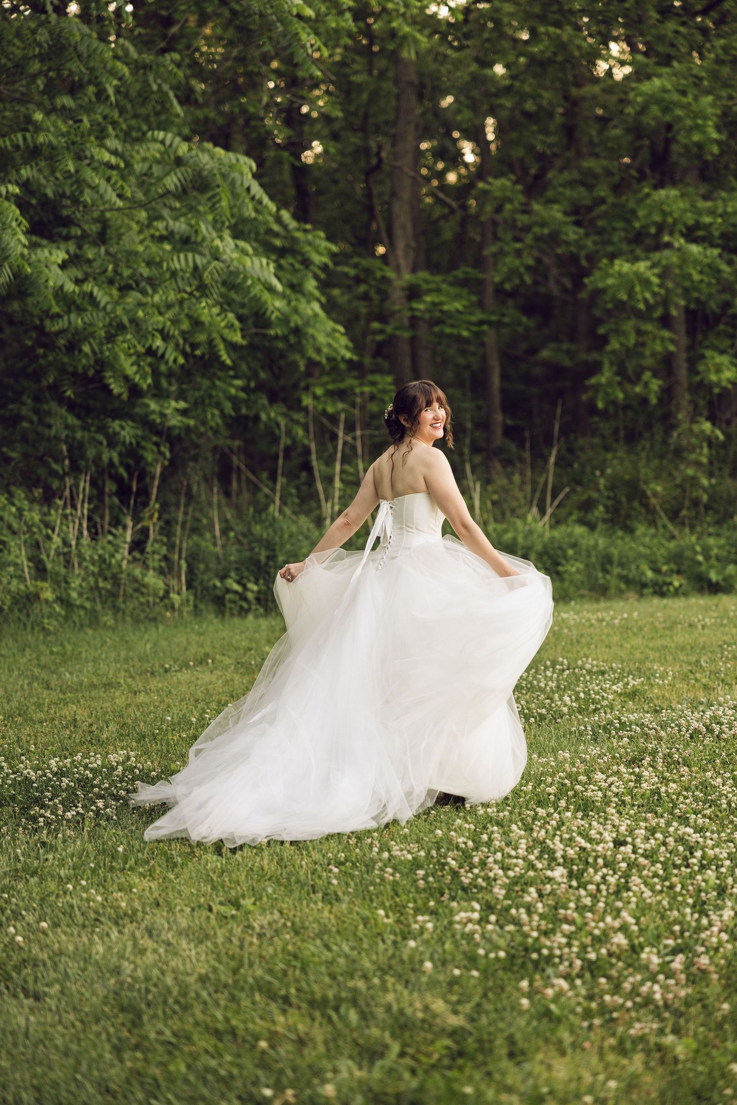 Bride in a white tulle wedding gown twirls joyfully across a green clover-filled lawn with a forest backdrop.