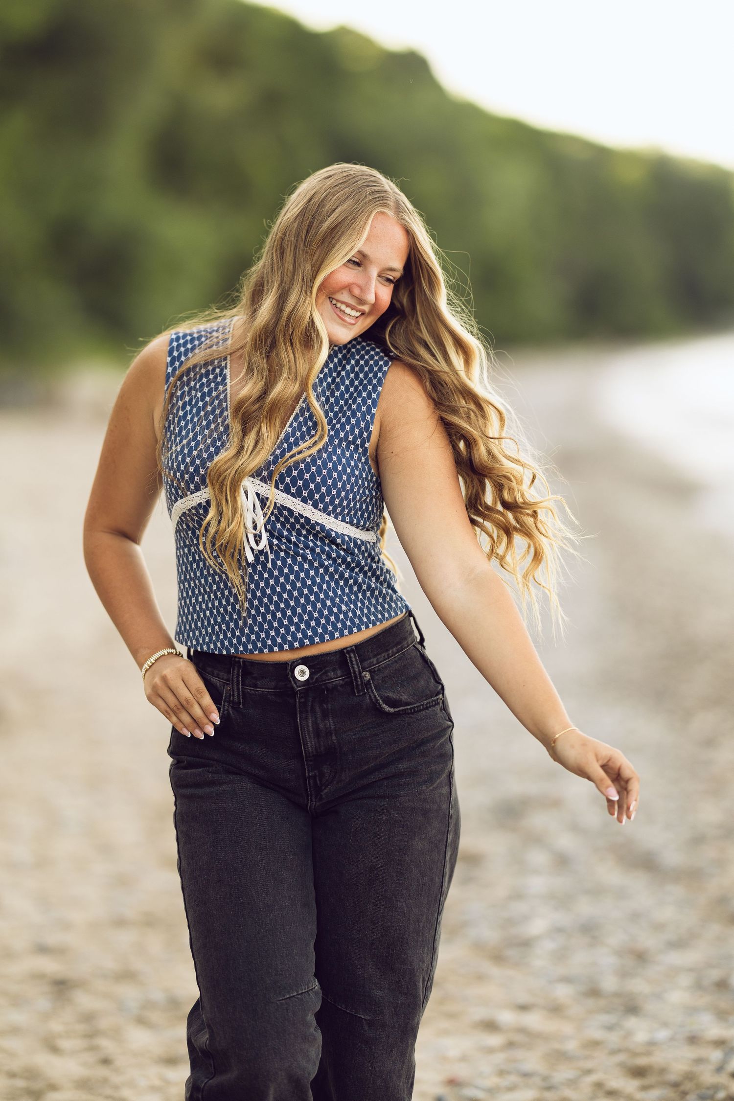 Laughing young woman with long wavy blonde hair in a blue patterned top and black jeans walks along a pebbly lakeshore.