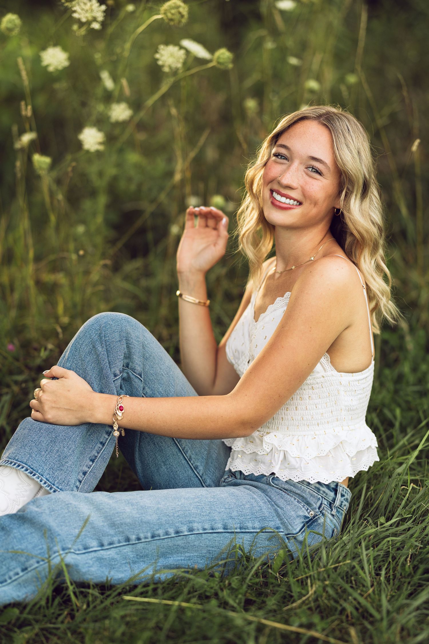 A smiling young woman with wavy blonde hair sits in tall green grass wearing a white ruffled top and jeans in golden light.