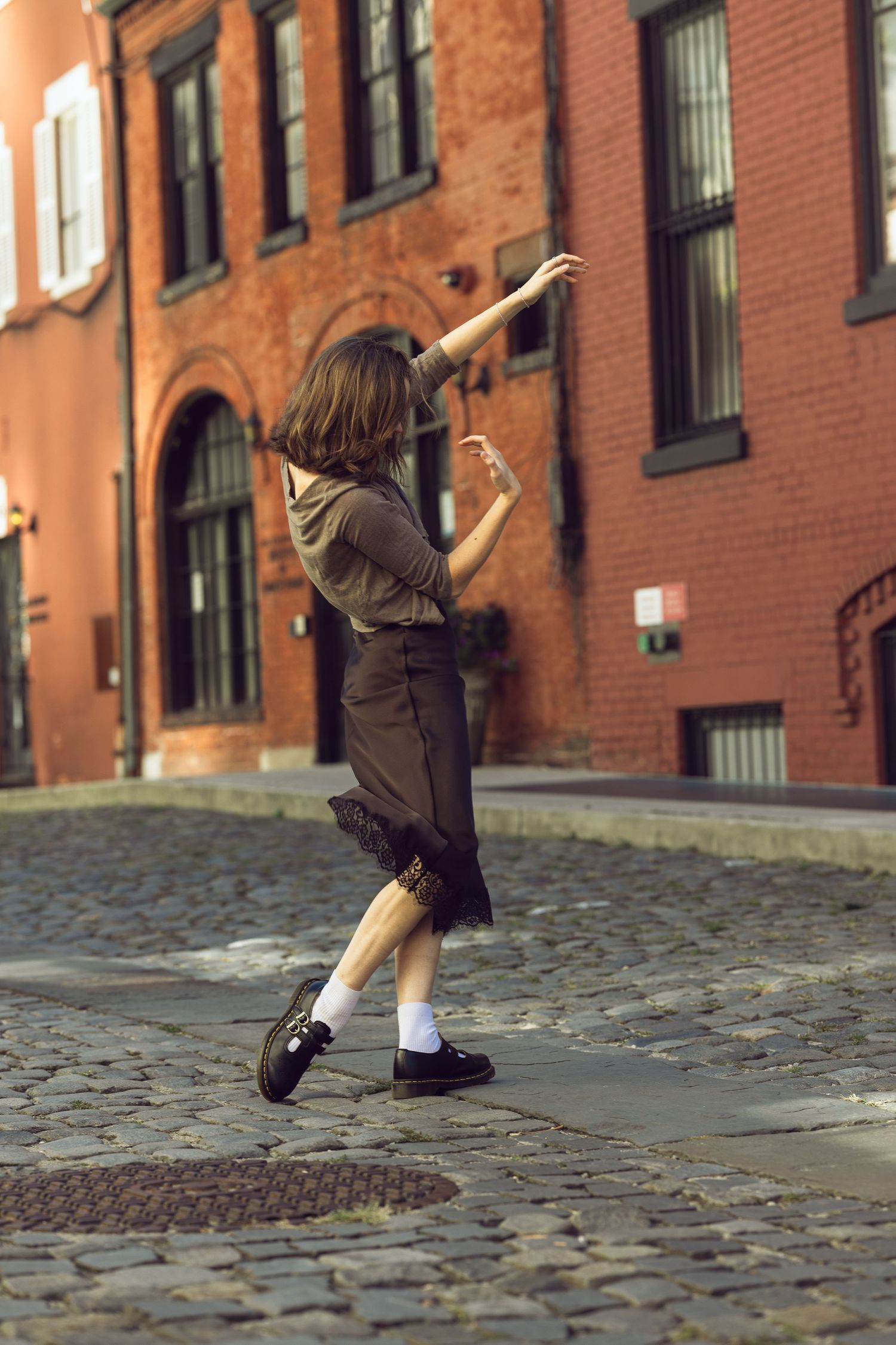 Woman in a brown top and skirt strikes a graceful dance pose on a cobblestone street in front of red brick buildings.