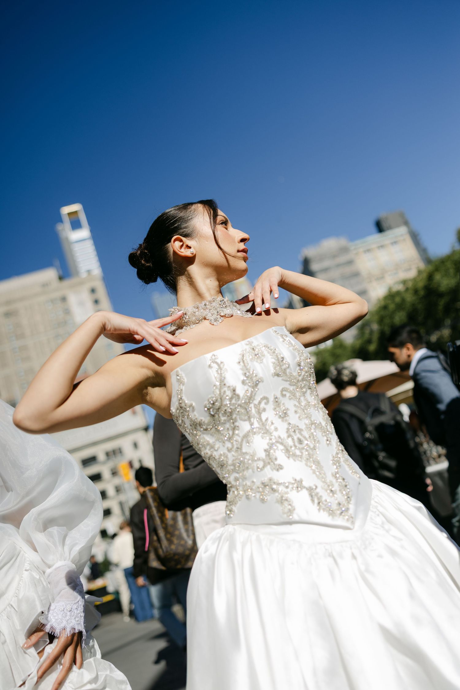 Bride in a beaded strapless satin wedding gown strikes a graceful pose against a blue sky and city buildings.