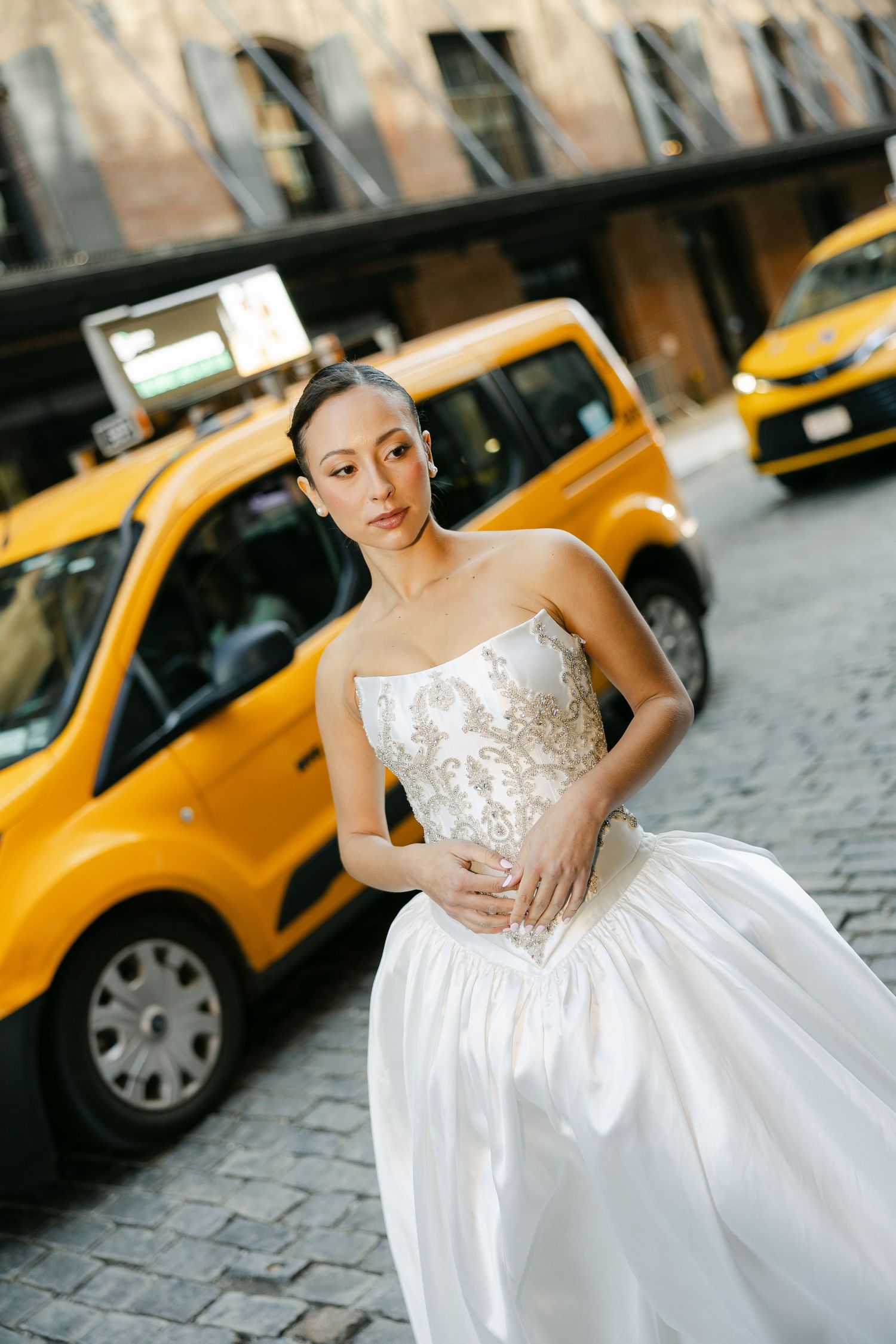 Bride in a strapless satin gown with beaded bodice poses on a cobblestone street beside yellow New York taxis.