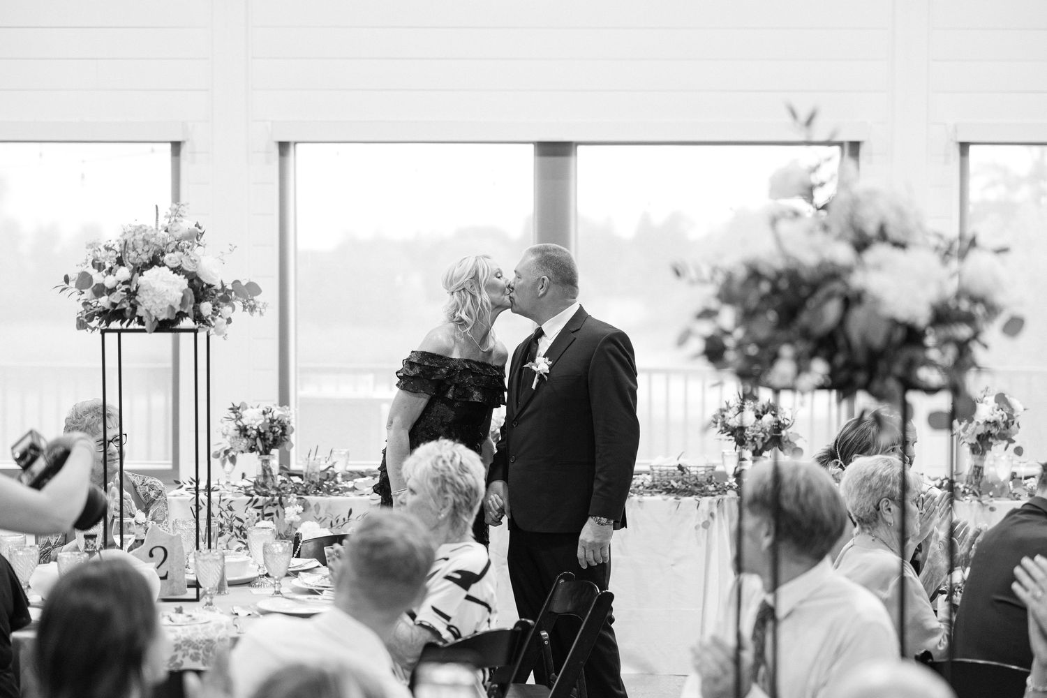 A black and white photo of a couple sharing a kiss beside reception tables with tall floral centerpieces as guests look on.
