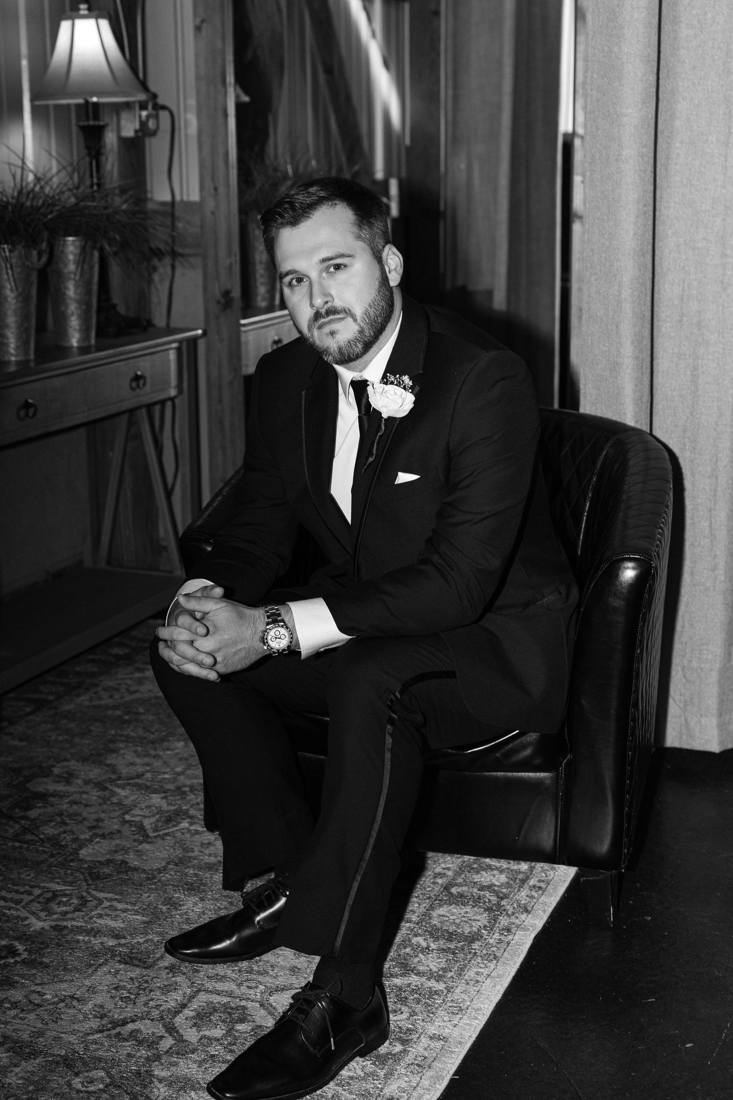 Black and white portrait of a bearded groom in a suit and boutonniere sitting in a leather chair with hands clasped.