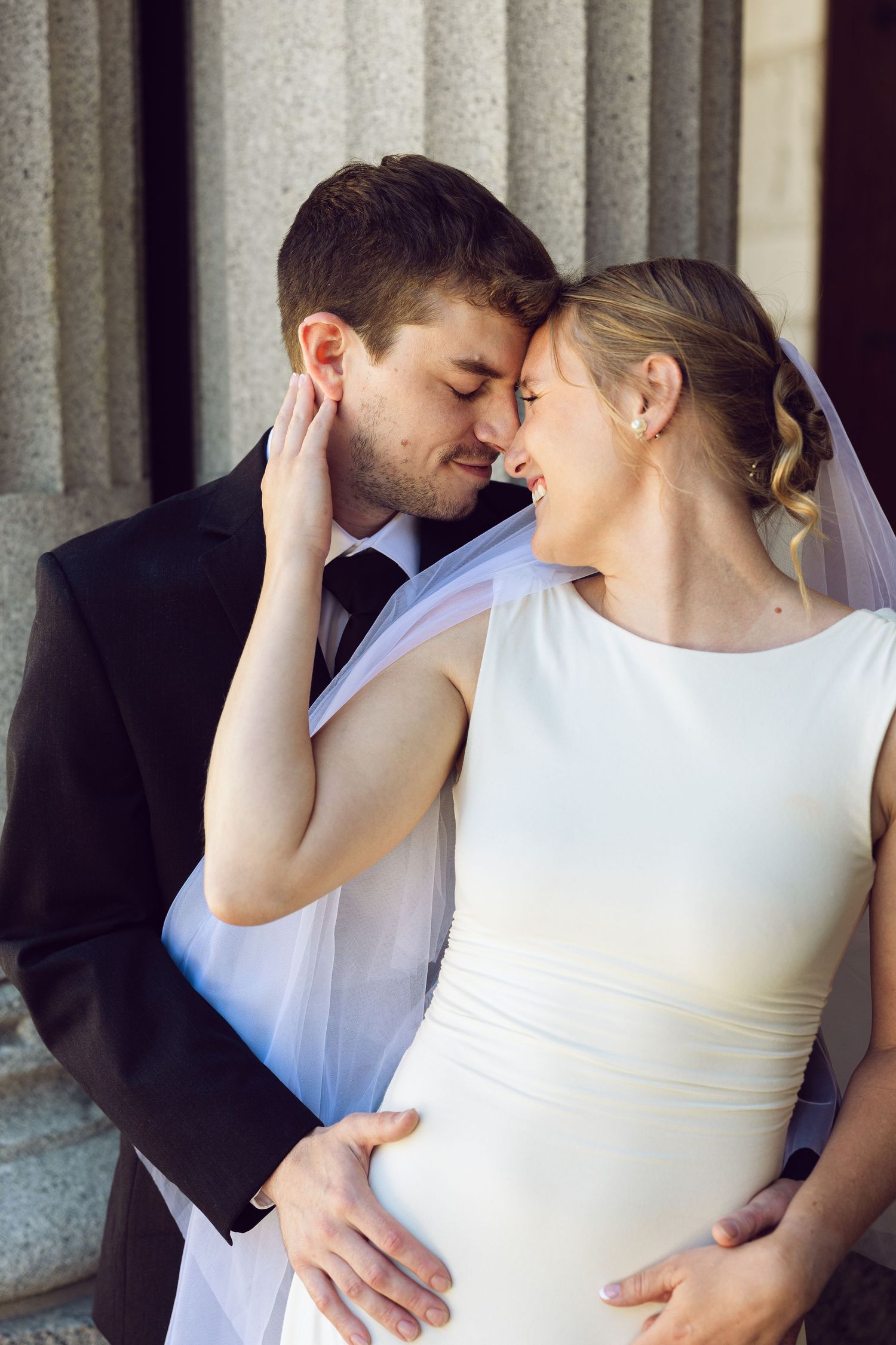 A bride and groom touch foreheads in a tender embrace in front of stone columns, her veil draped over his arm.