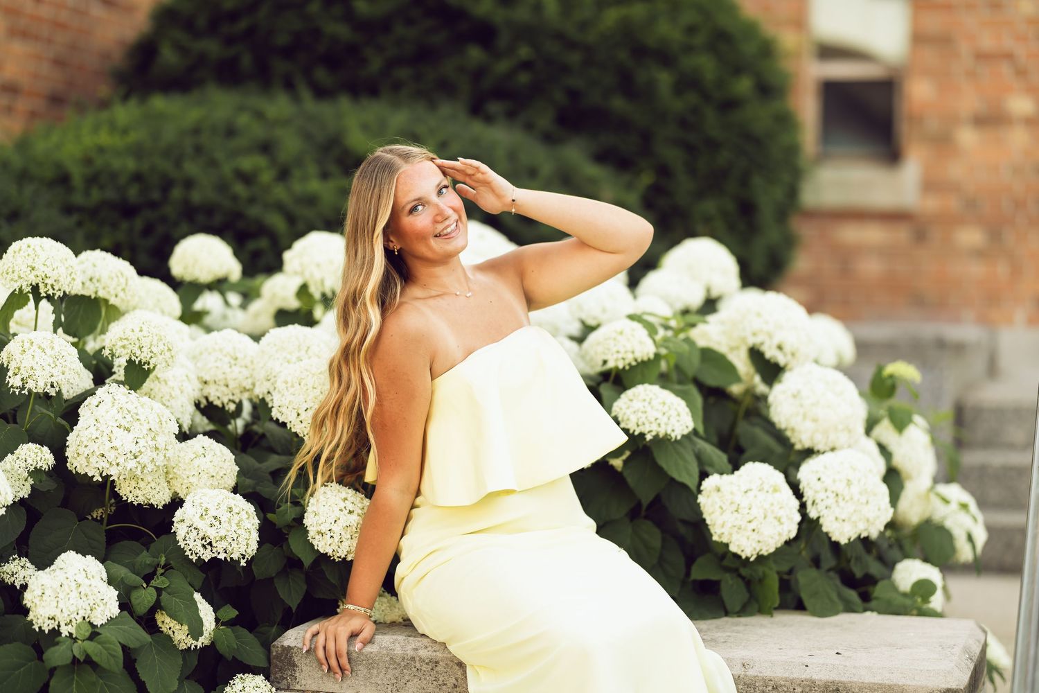 Young woman in a pale yellow strapless dress sits beside white hydrangeas with a brick building behind her, hand in hair.