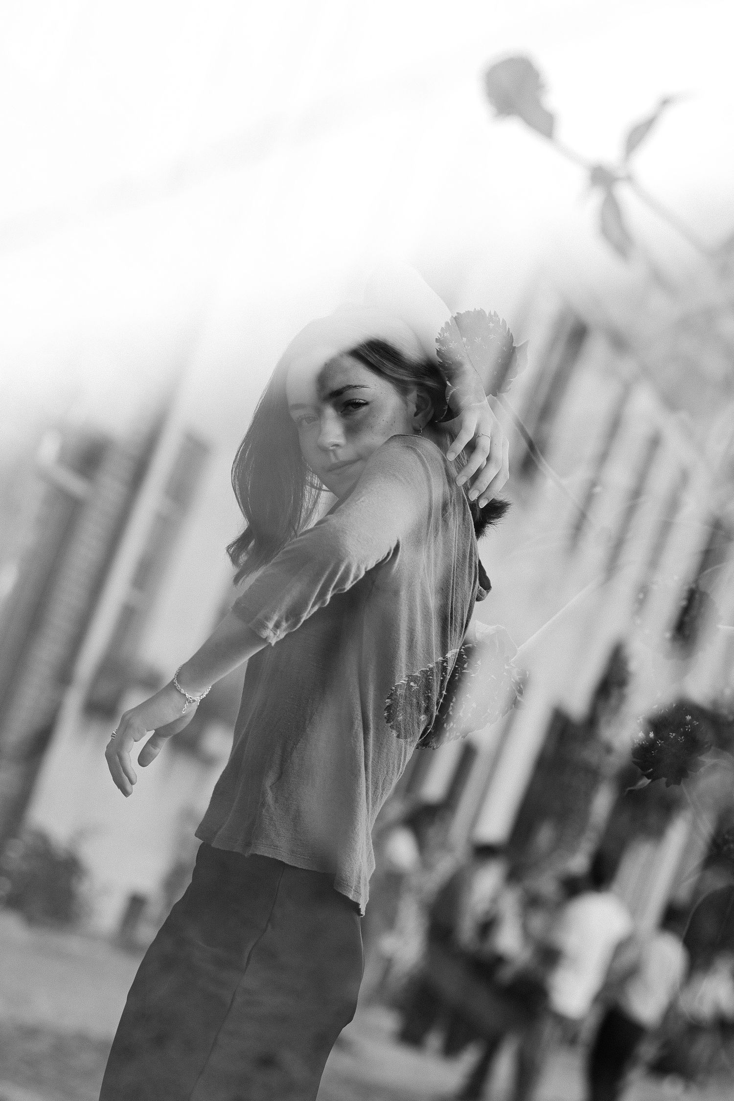 Black and white dreamy portrait of a woman posing with arms raised on a city street with softly blurred pedestrians behind her.