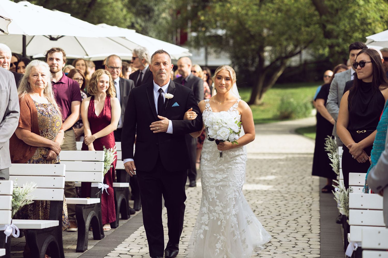 Bride in a lace gown holding a white bouquet walks down the outdoor wedding aisle arm in arm with a man in a dark suit.