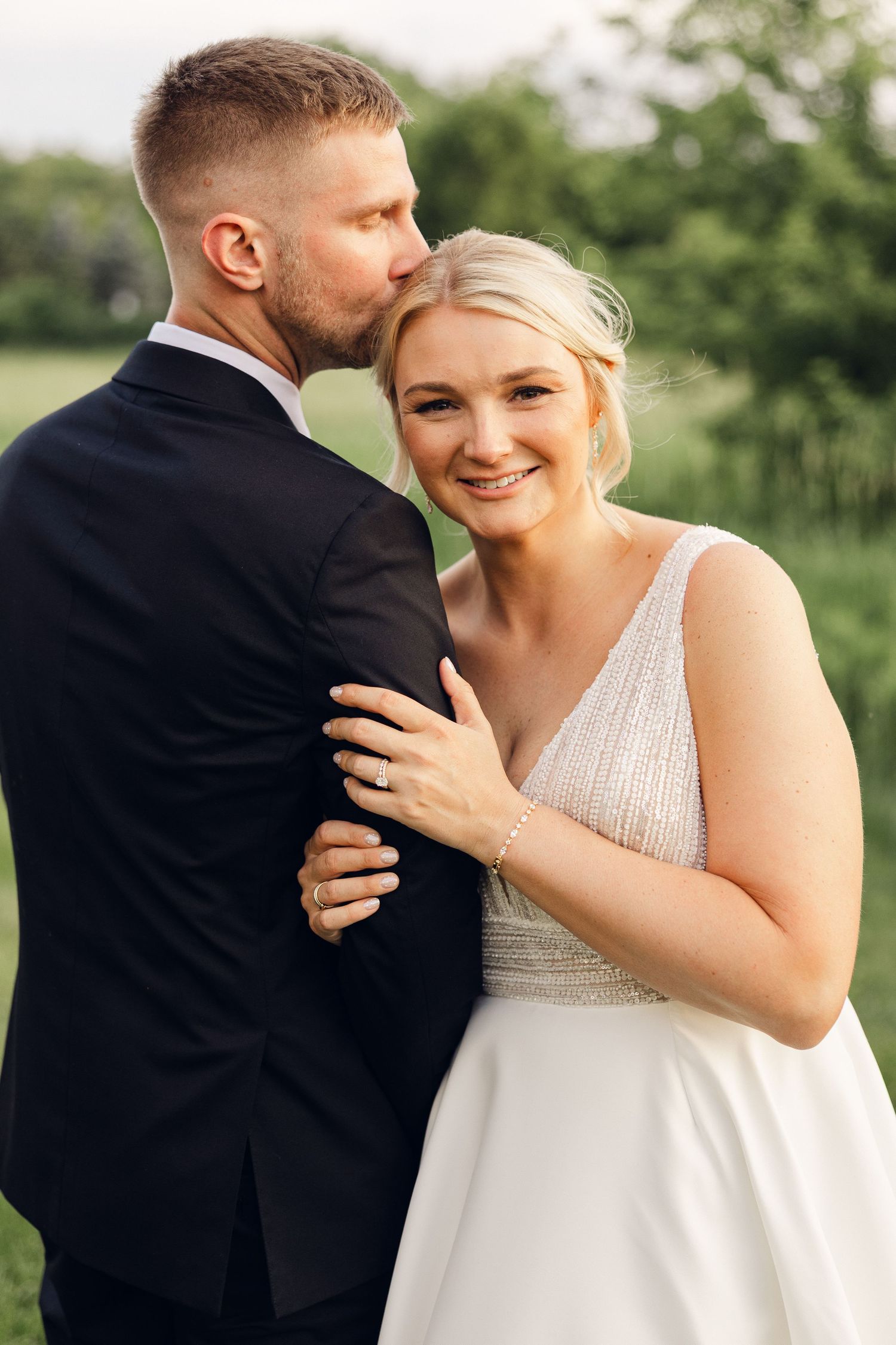 Groom kisses the smiling blonde bride on the forehead as she rests her hand on his arm in a green outdoor setting.