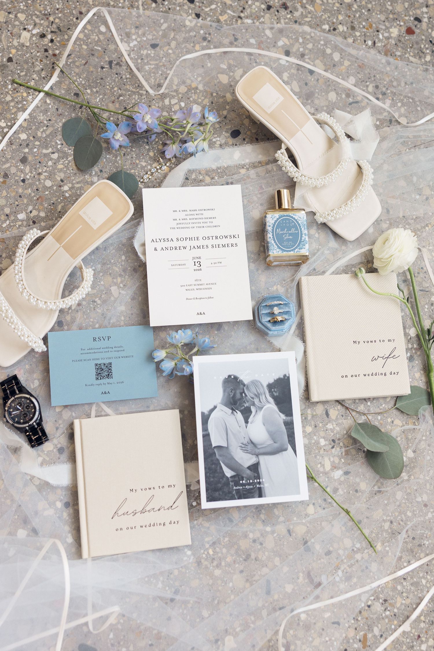 A wedding flat lay with an invitation, RSVP card, husband and wife keepsake books, pearl sandals, perfume, ring box, and a couple's photo.