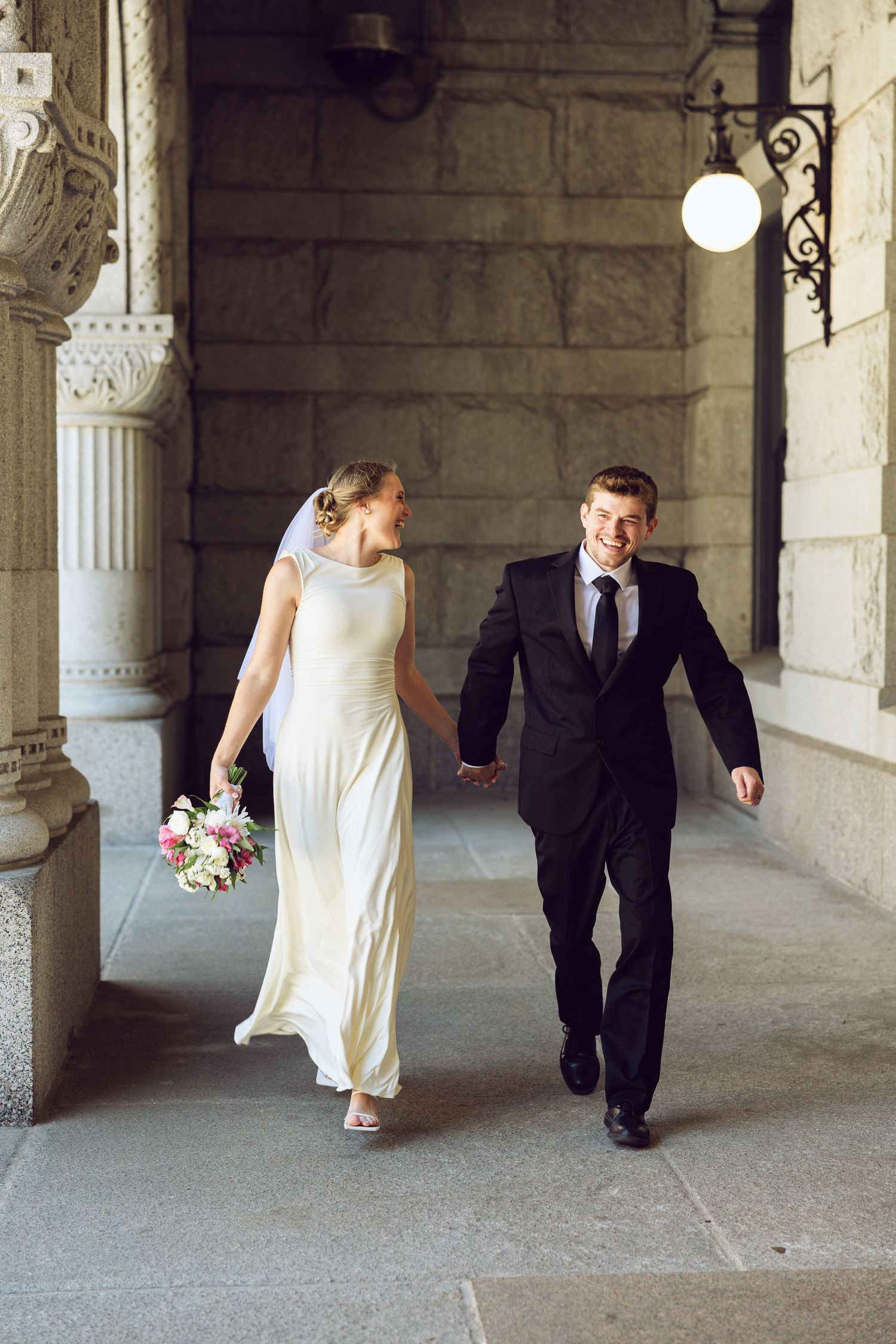 A laughing bride and groom hold hands and walk through a stone archway, her bouquet in hand and her gown flowing.