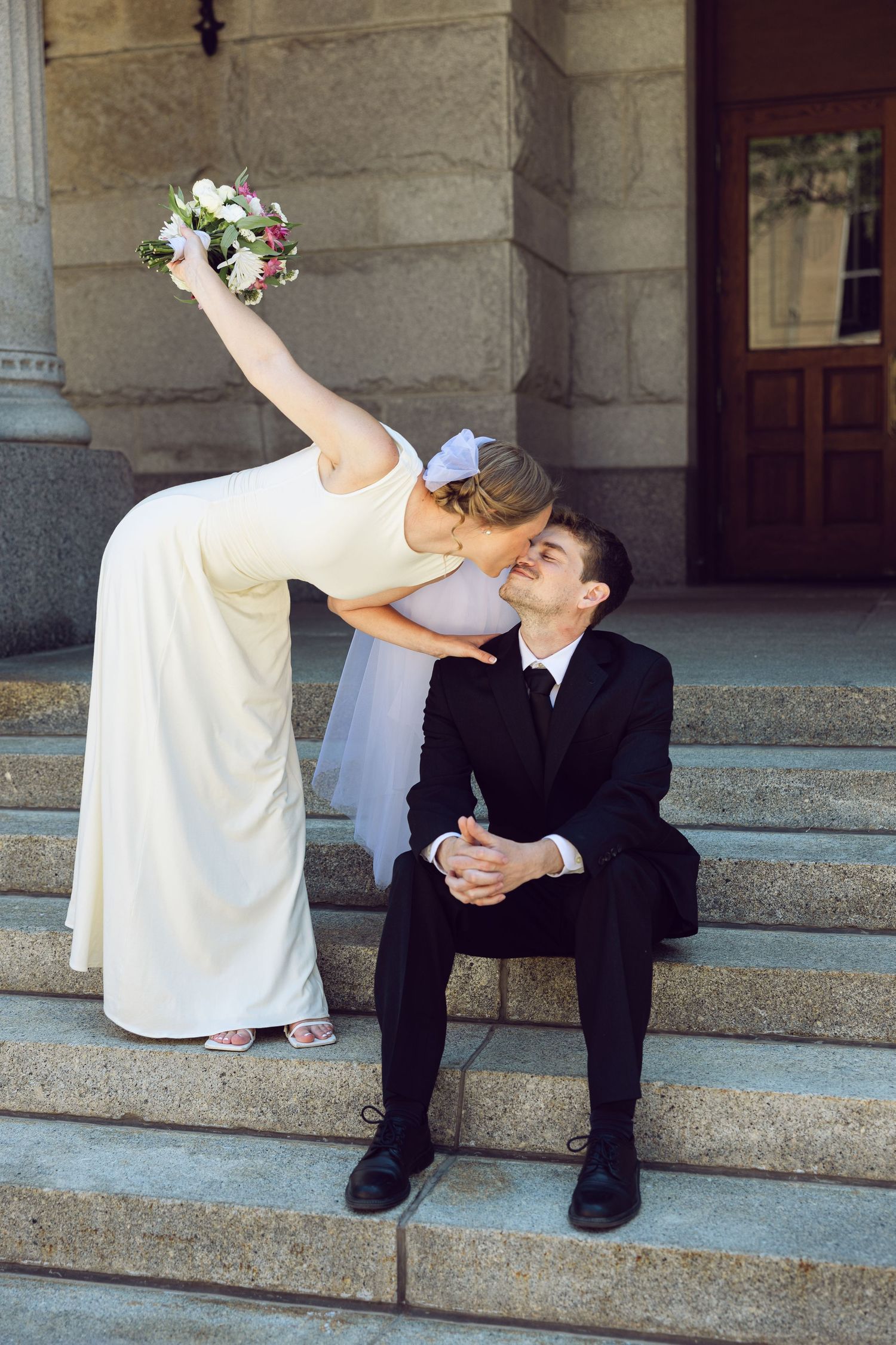 A bride in a white gown leans down to kiss her seated groom on stone steps while lifting her bouquet high in the air.