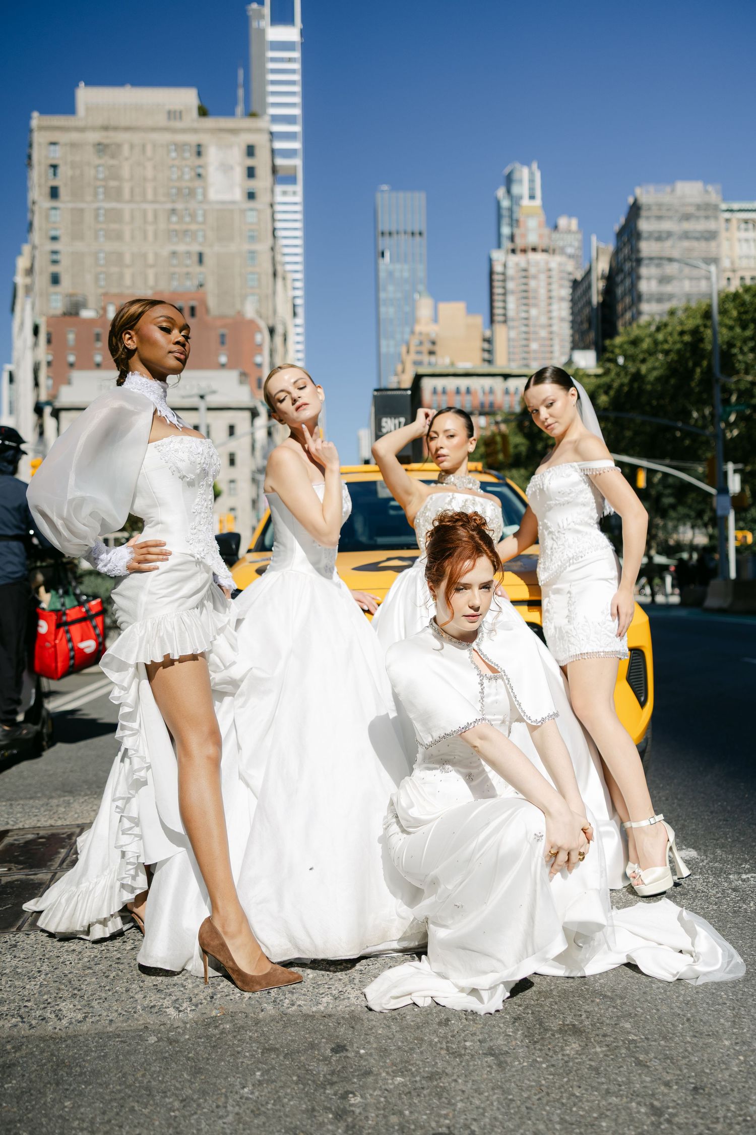 Four models in white bridal gowns pose in front of a yellow taxi on a sunny New York City street.