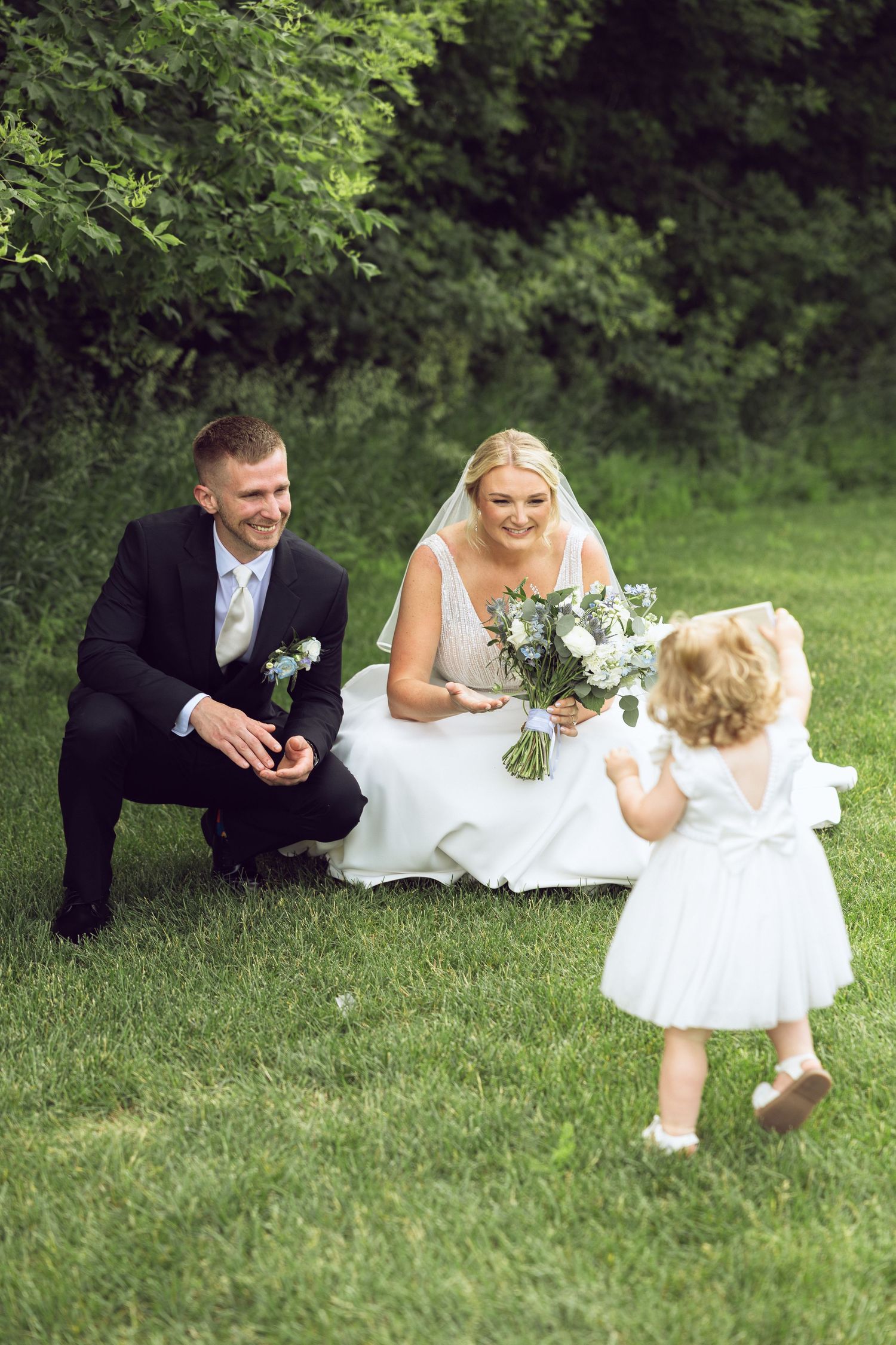Bride and groom crouch on the grass with a bouquet as their toddler flower girl in a white dress walks toward them.