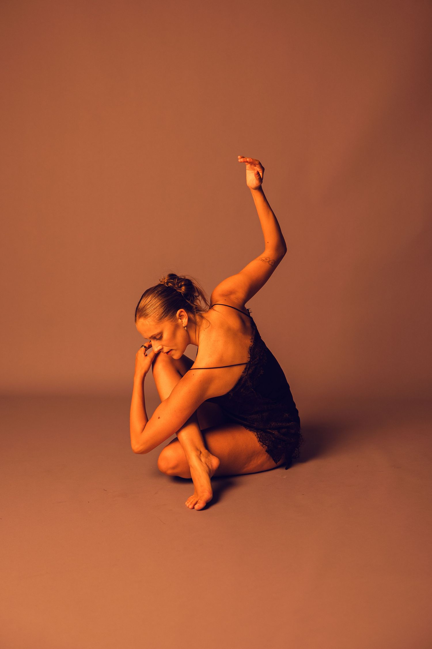 Dancer in a dark dress sits in a folded pose with one arm raised high against a warm amber studio background.