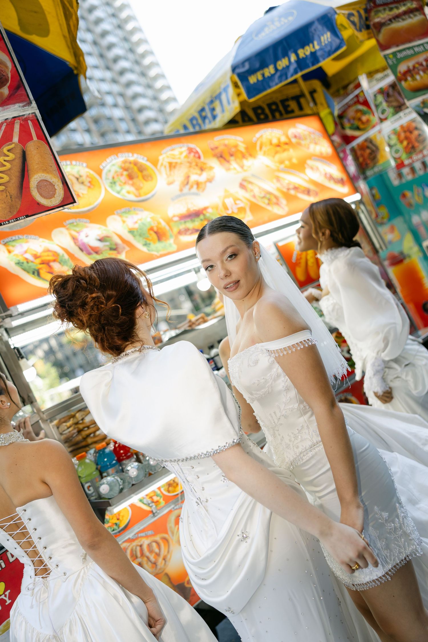 Brides in assorted white wedding gowns and veils pose together in front of a colorful New York City street food cart.