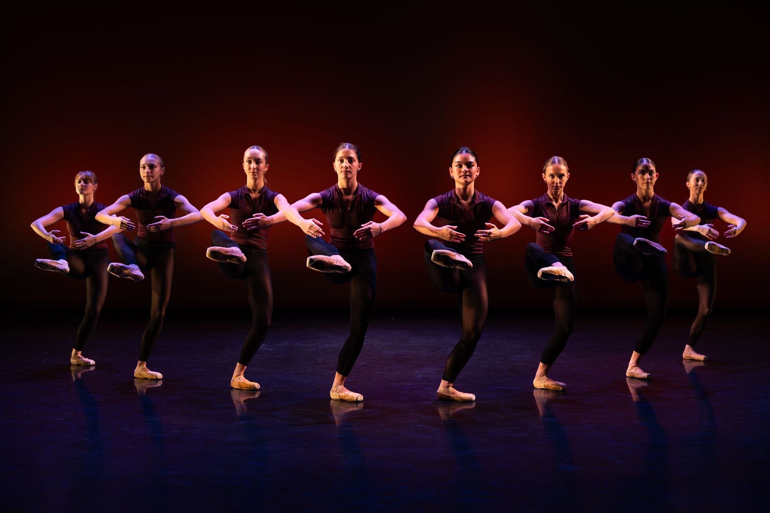 A line of dancers in black stand in a synchronized ballet pose on a dark stage lit with warm red and orange tones.