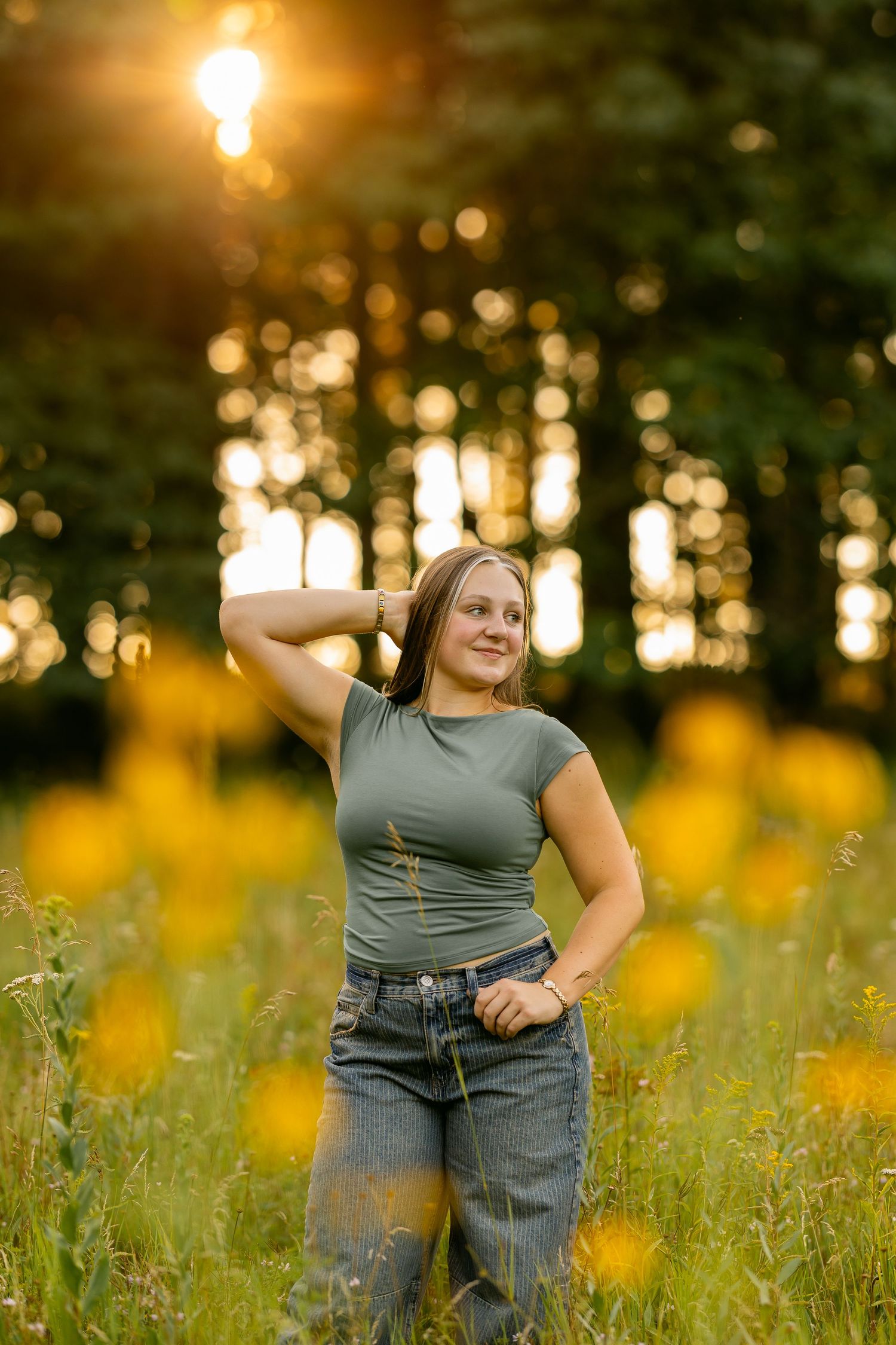 Young woman in a green top and jeans smiles with her hand in her hair in a yellow wildflower field at sunset.