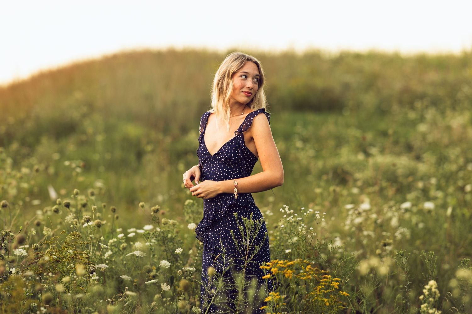 Blonde woman in a navy floral sundress stands in a sunlit wildflower meadow with rolling green hills in the background.