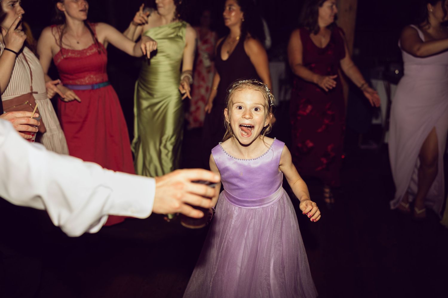 Smiling flower girl in a lavender tulle dress dances joyfully among wedding guests on the reception dance floor.