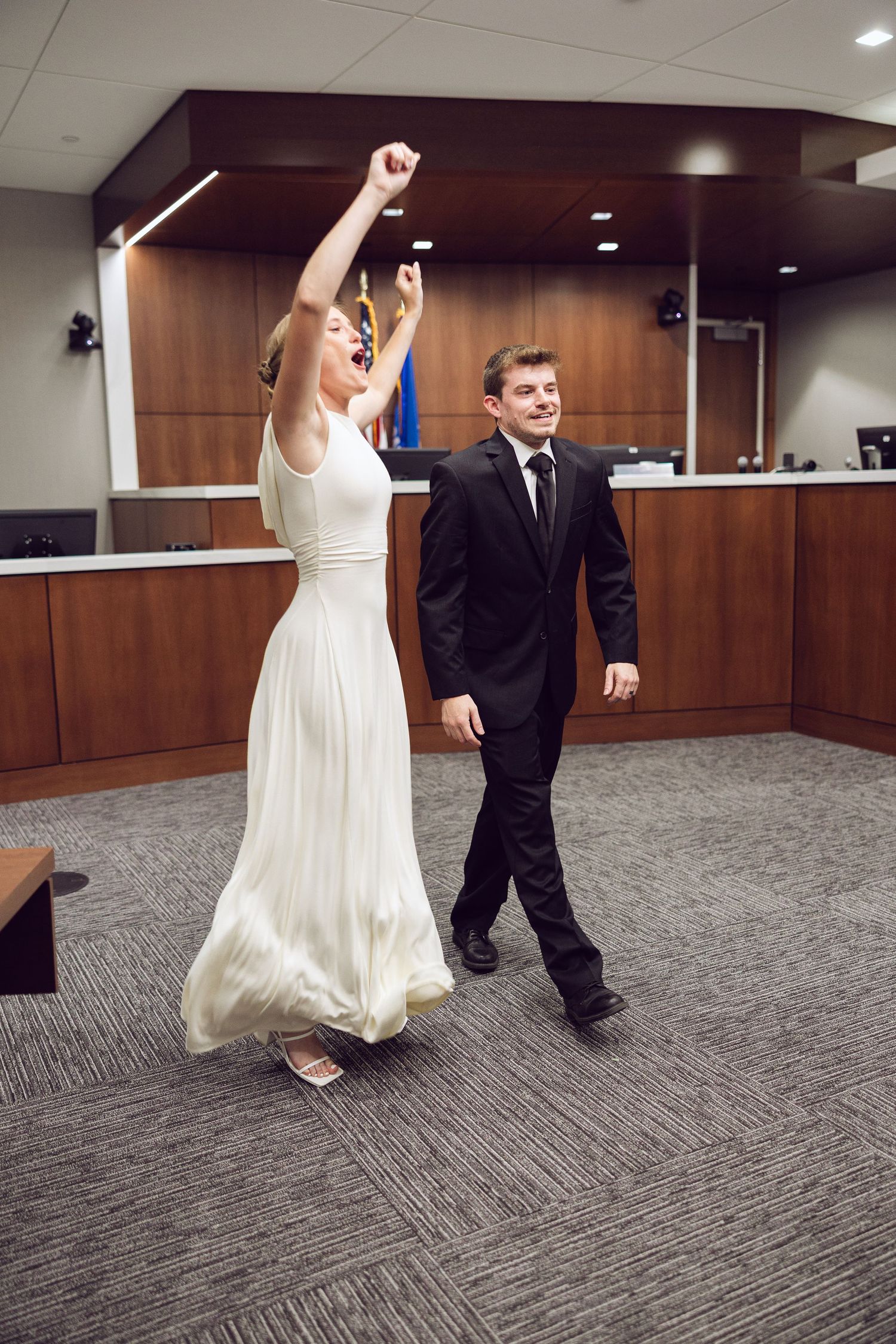 Bride raises her arms in celebration while the groom smiles and walks beside her in a courthouse after their wedding.