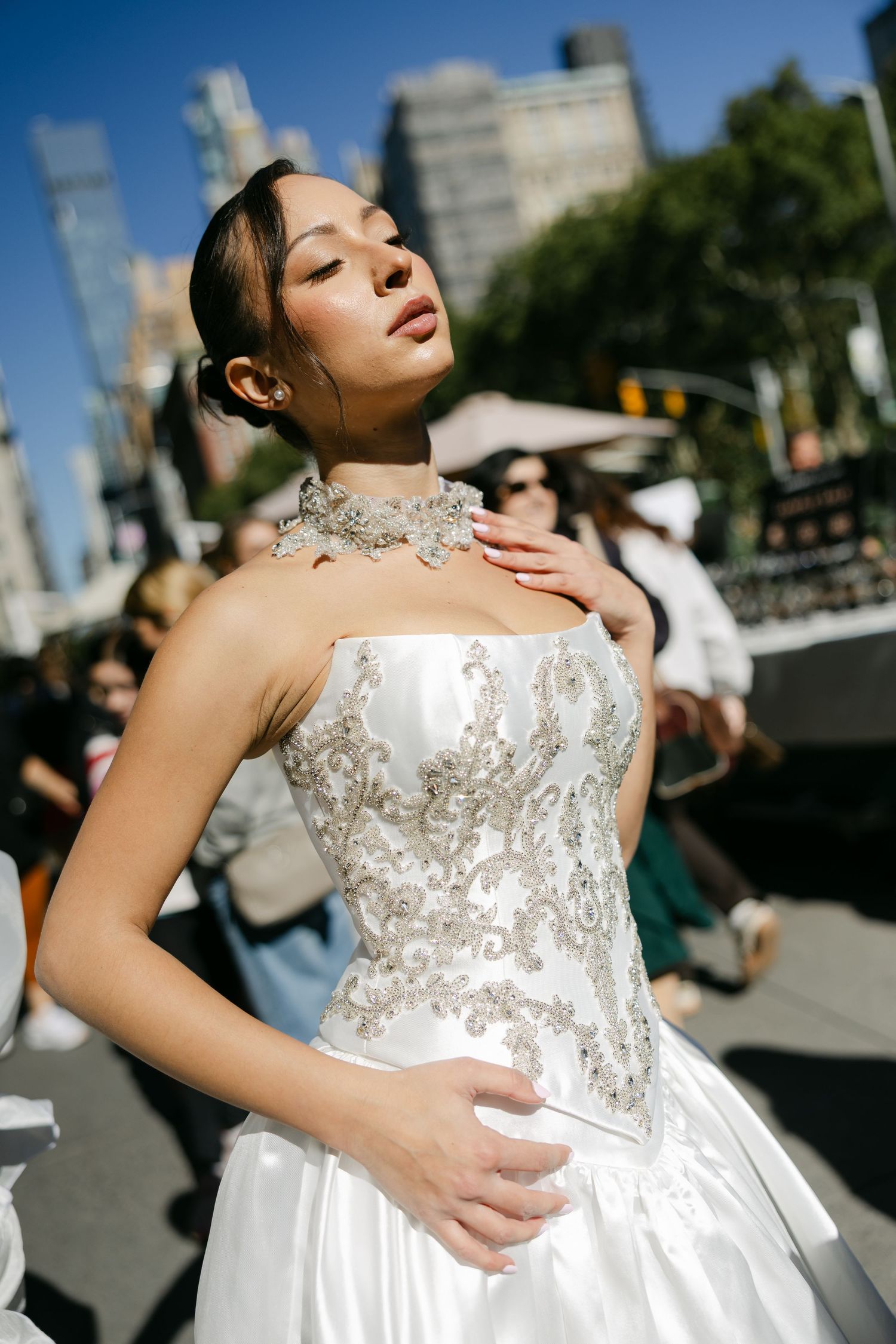 A model in a strapless white bridal gown with silver beaded embroidery and a matching jeweled choker poses on a busy city street.