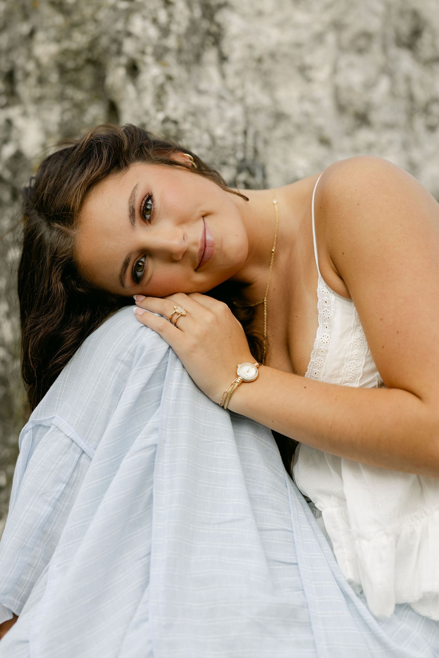 Young woman with long dark wavy hair in a white camisole smiles while resting her head on her hand against a stone wall.