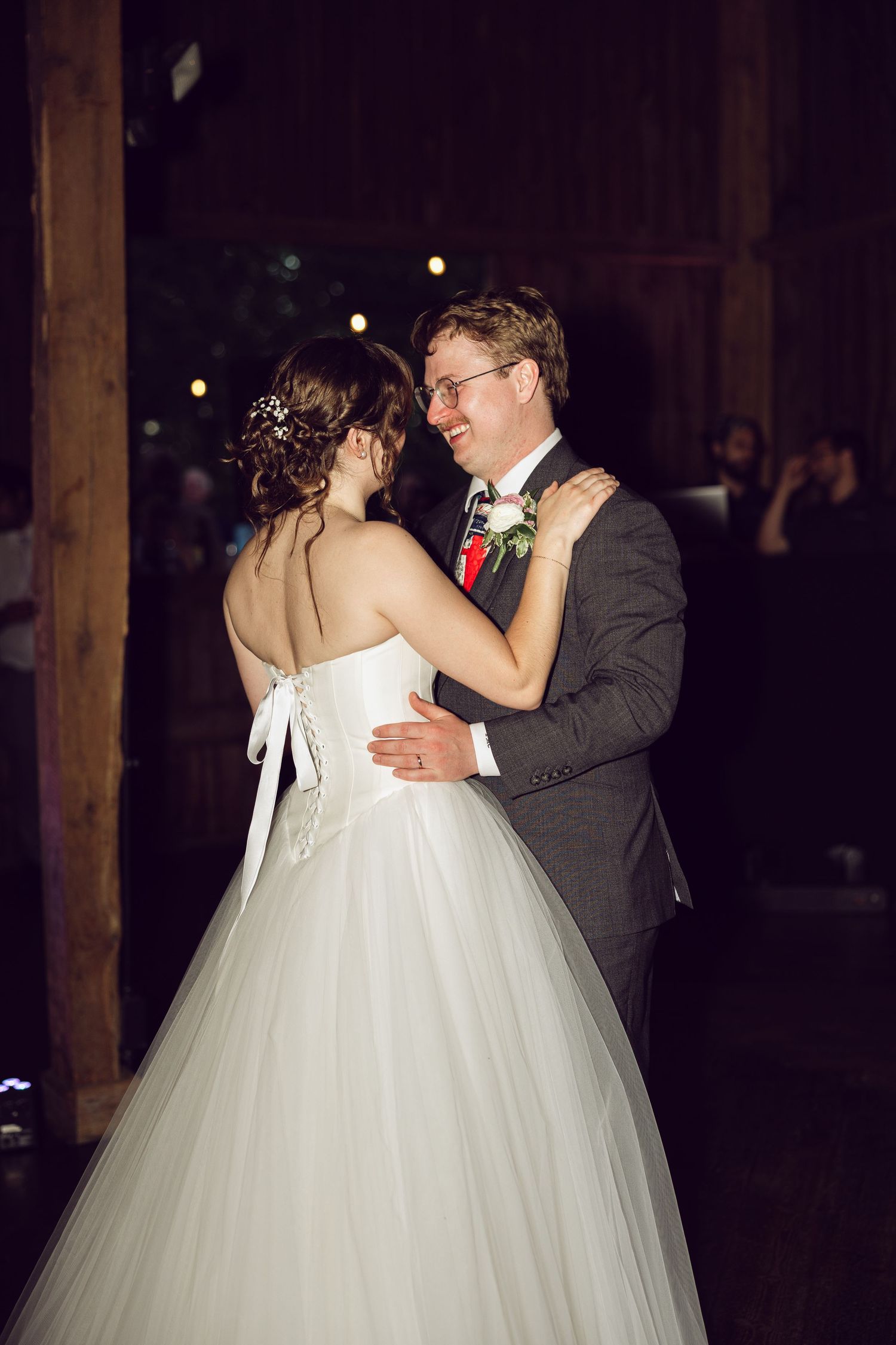 Bride in a white tulle gown with a floral hair accessory dances with a smiling groom in glasses at a rustic barn wedding.