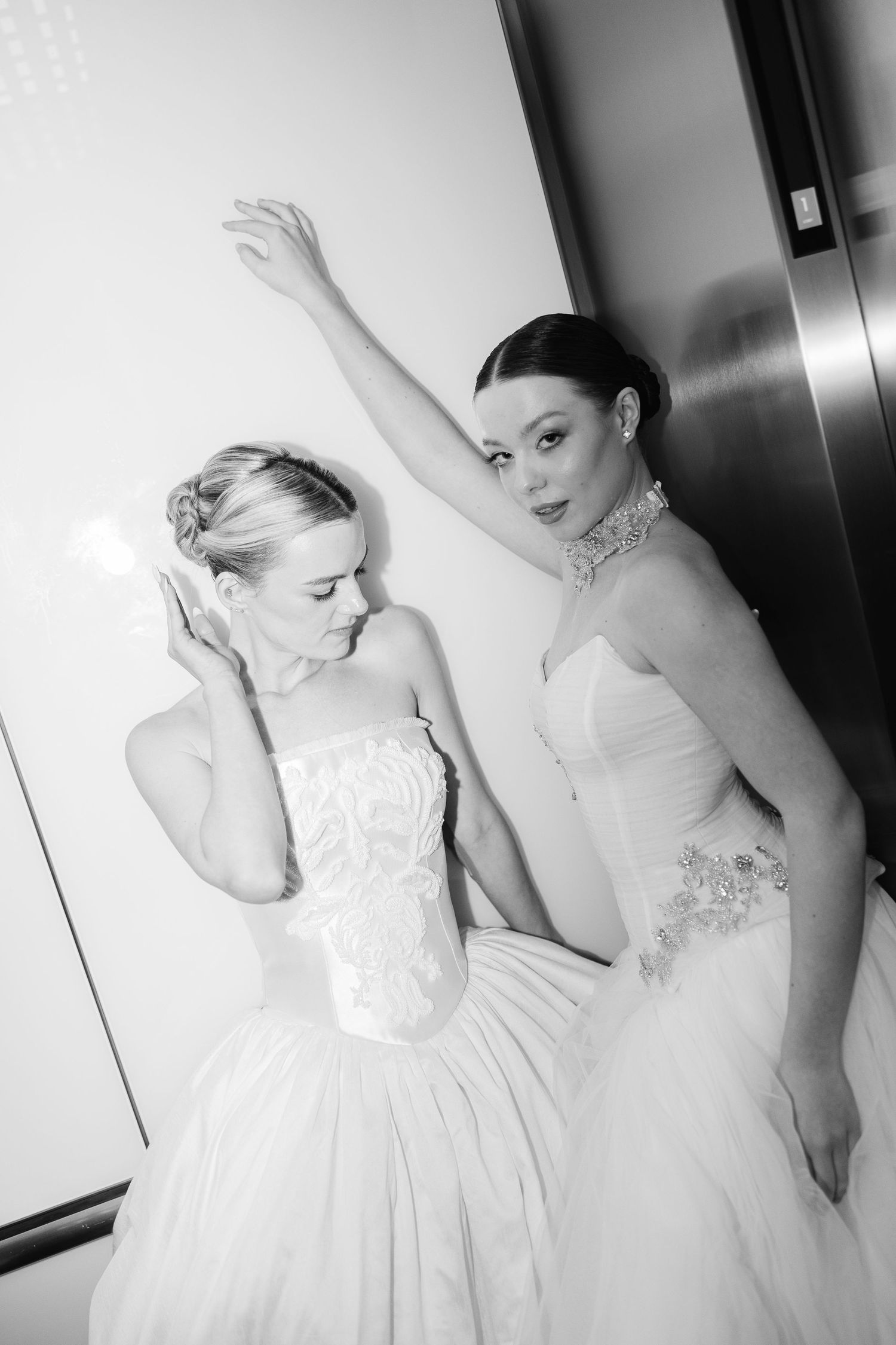 Two brides in strapless embellished tulle wedding gowns pose dramatically in a black and white editorial photo.