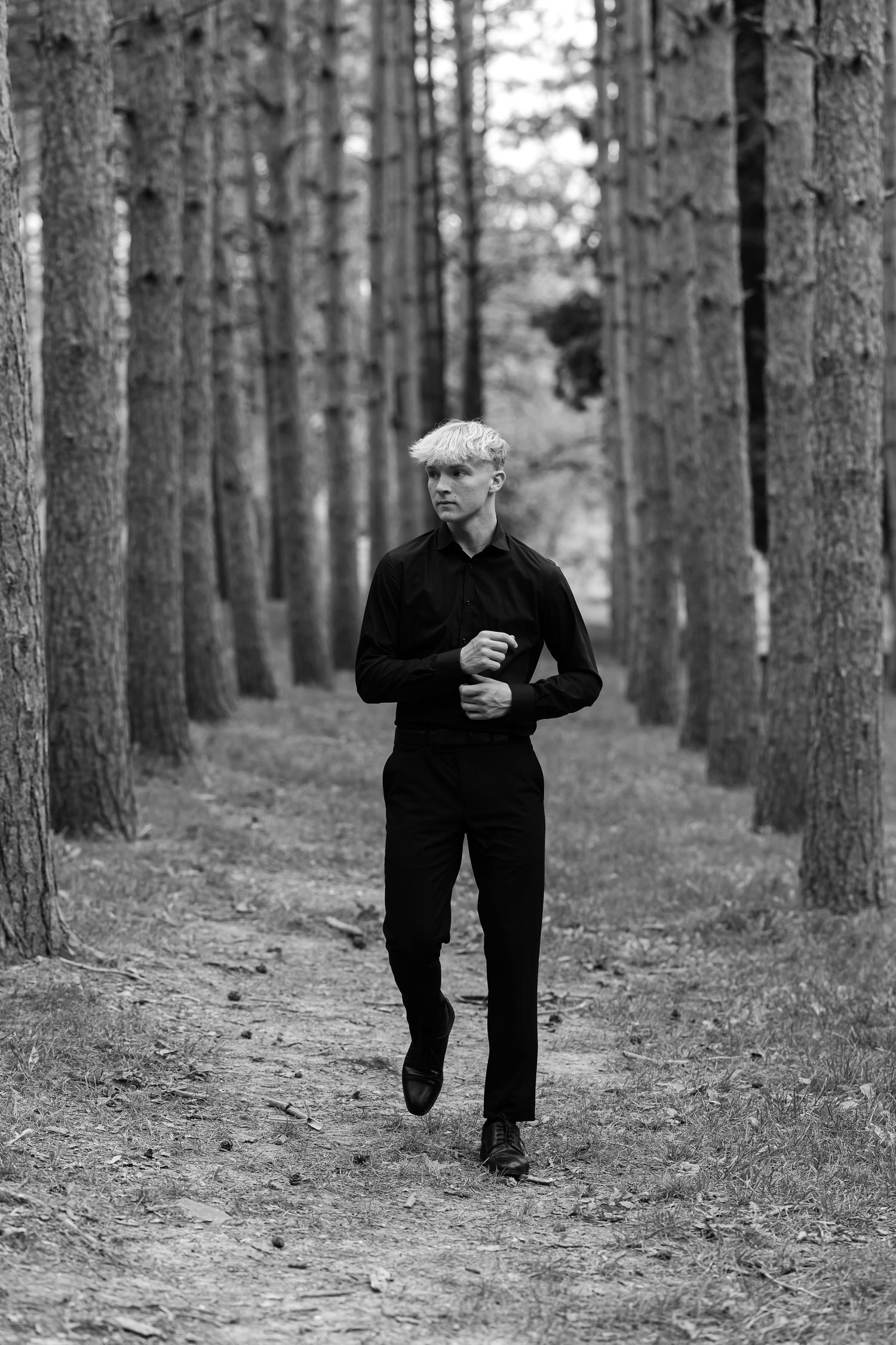 Black and white portrait of a blond young man in dark clothing walking down a path lined with tall pine trees.