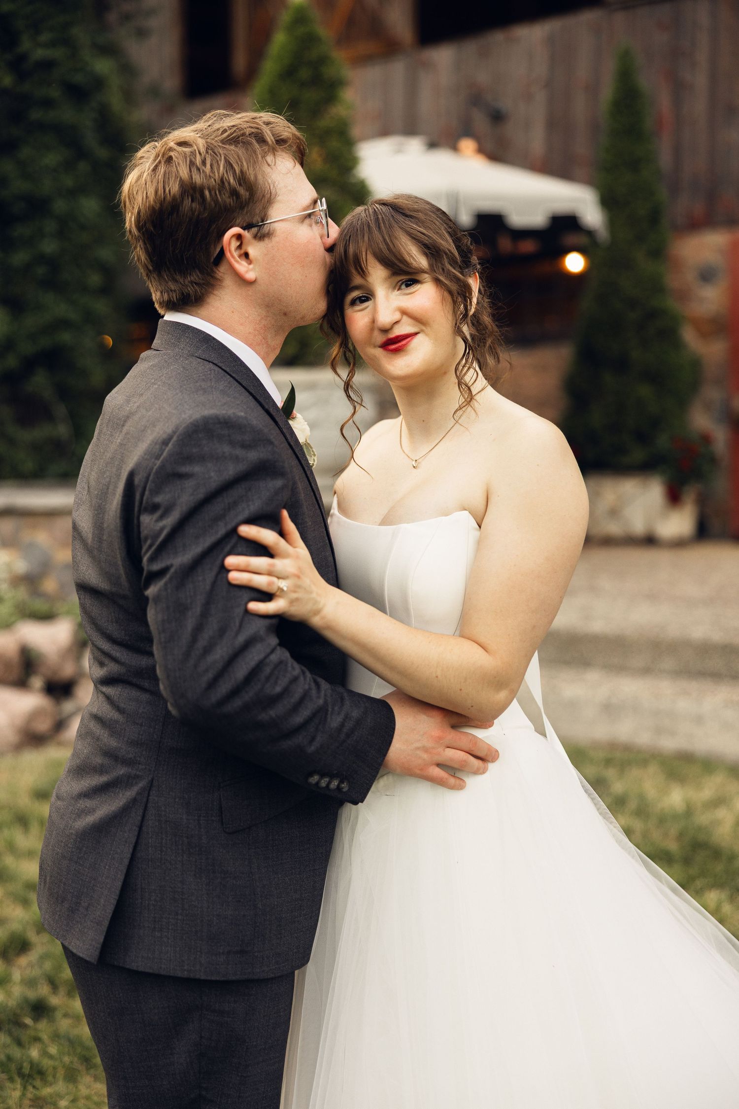 Groom in a gray suit kisses his smiling bride on the forehead outside a rustic barn wedding venue.
