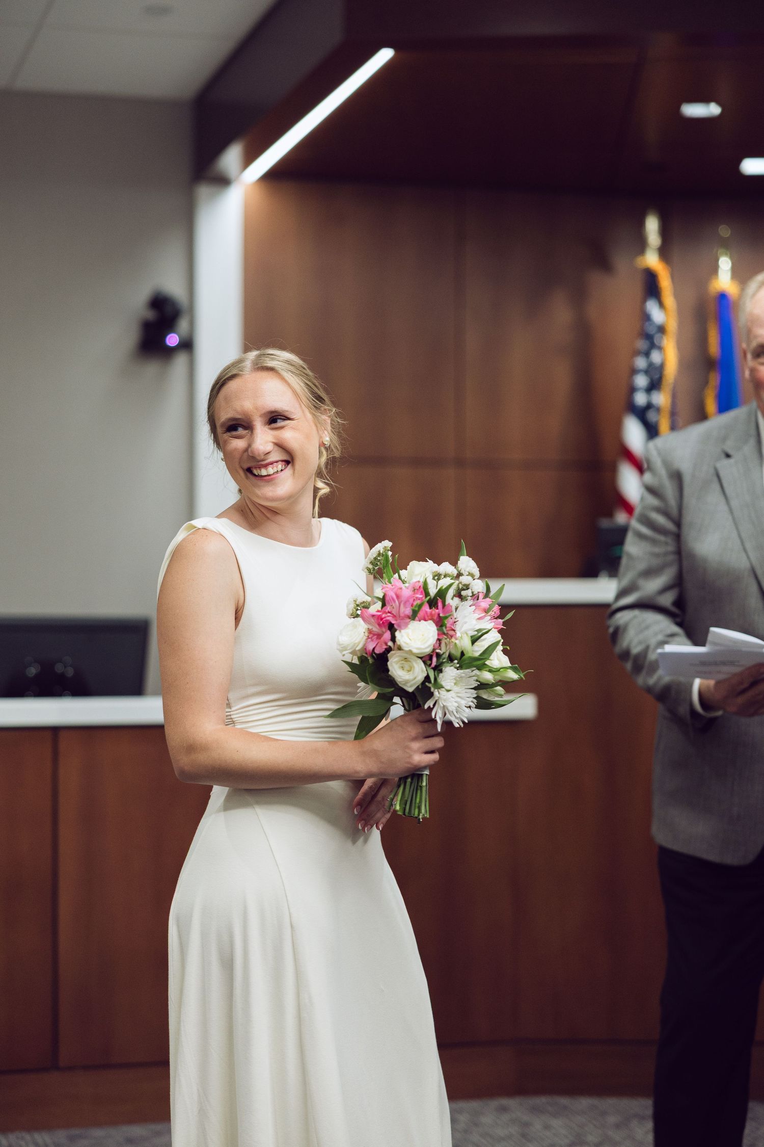 Smiling bride in a white sleeveless dress holds a pink and white bouquet during her courthouse wedding ceremony.
