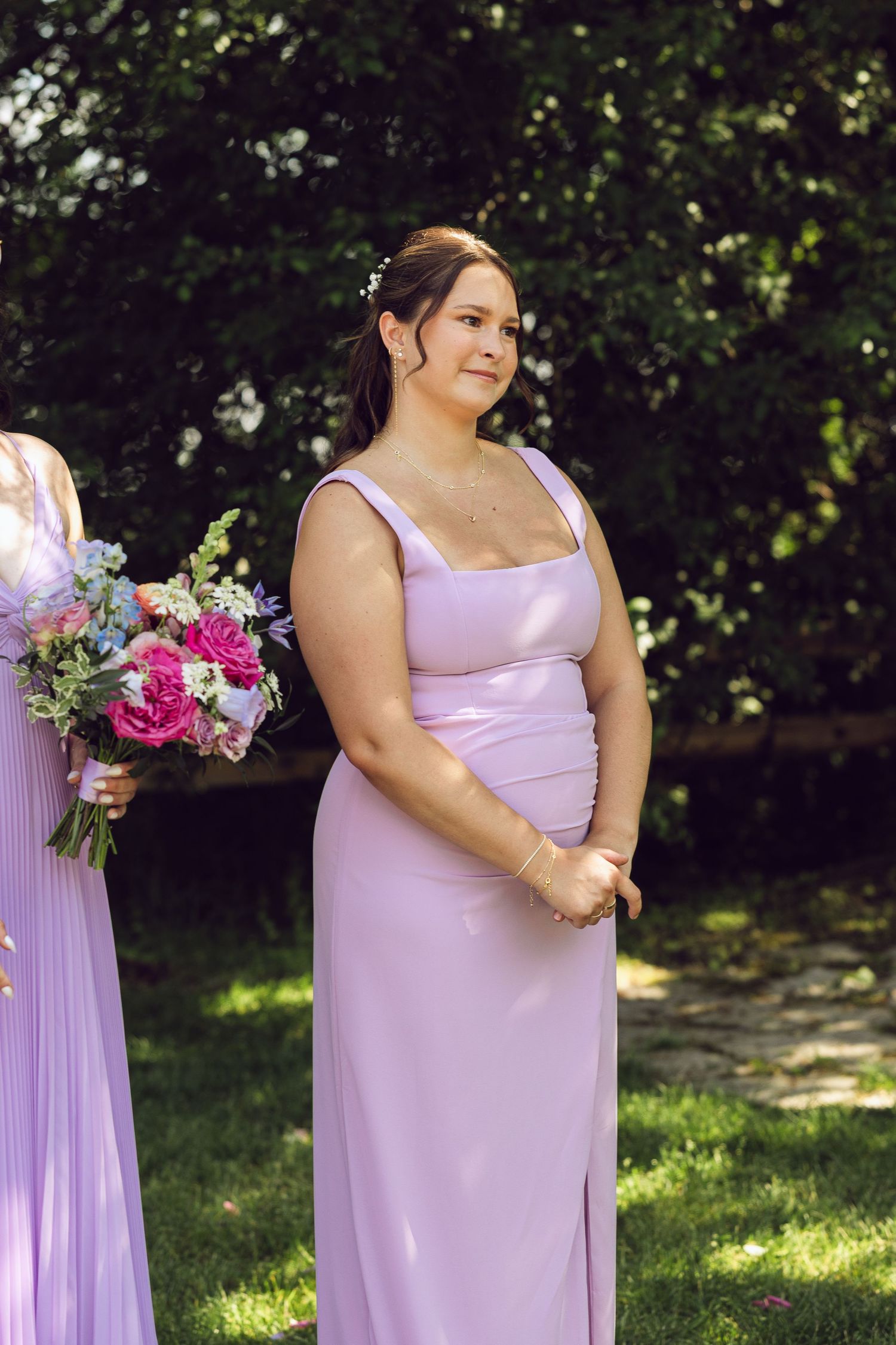 Bridesmaid in a lilac gown stands smiling at an outdoor wedding ceremony while another holds a pink and blue bouquet.