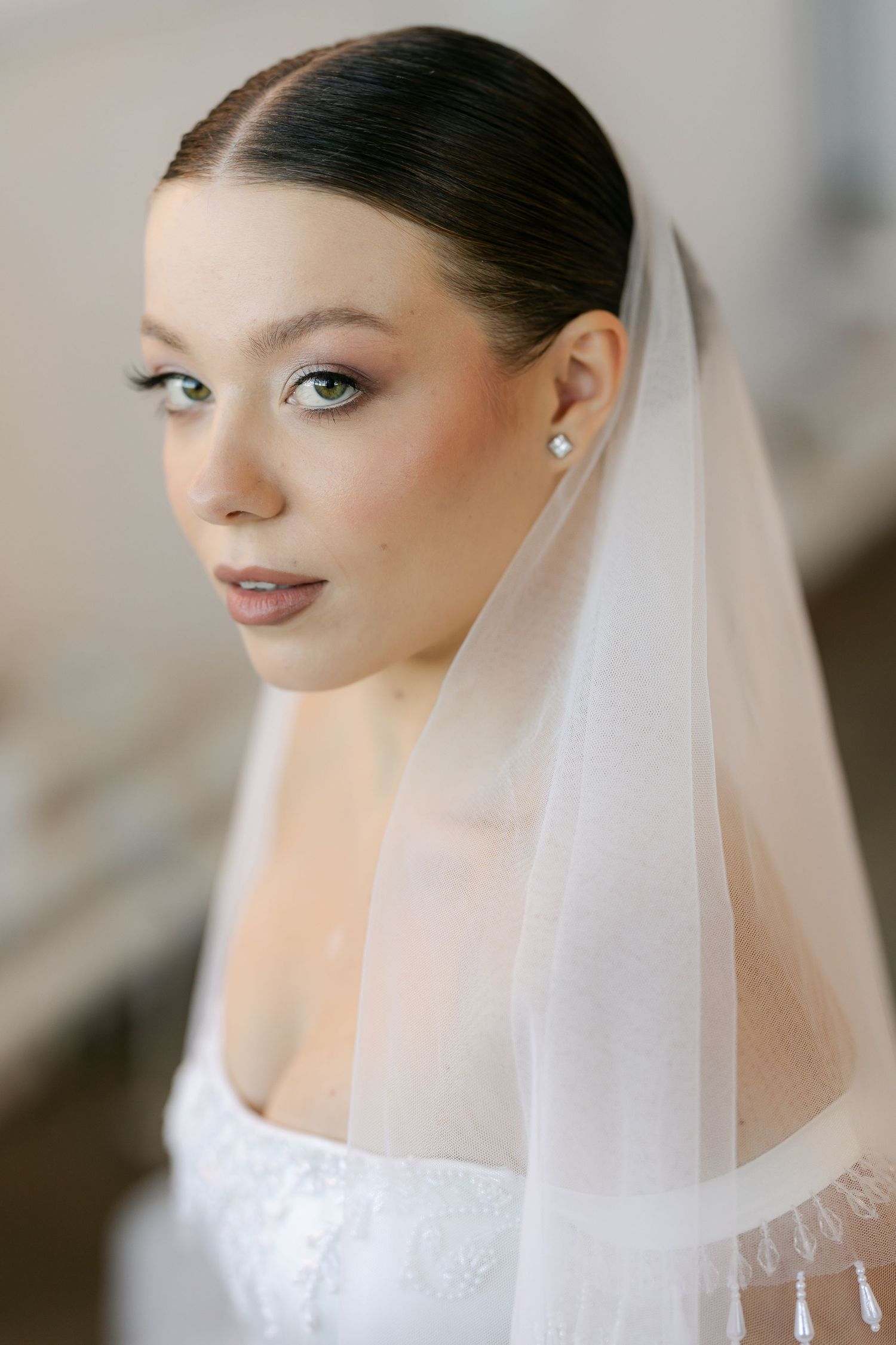 Close-up portrait of a bride with sleek dark hair, soft makeup, pearl earrings and a sheer tulle veil over a lace bodice.