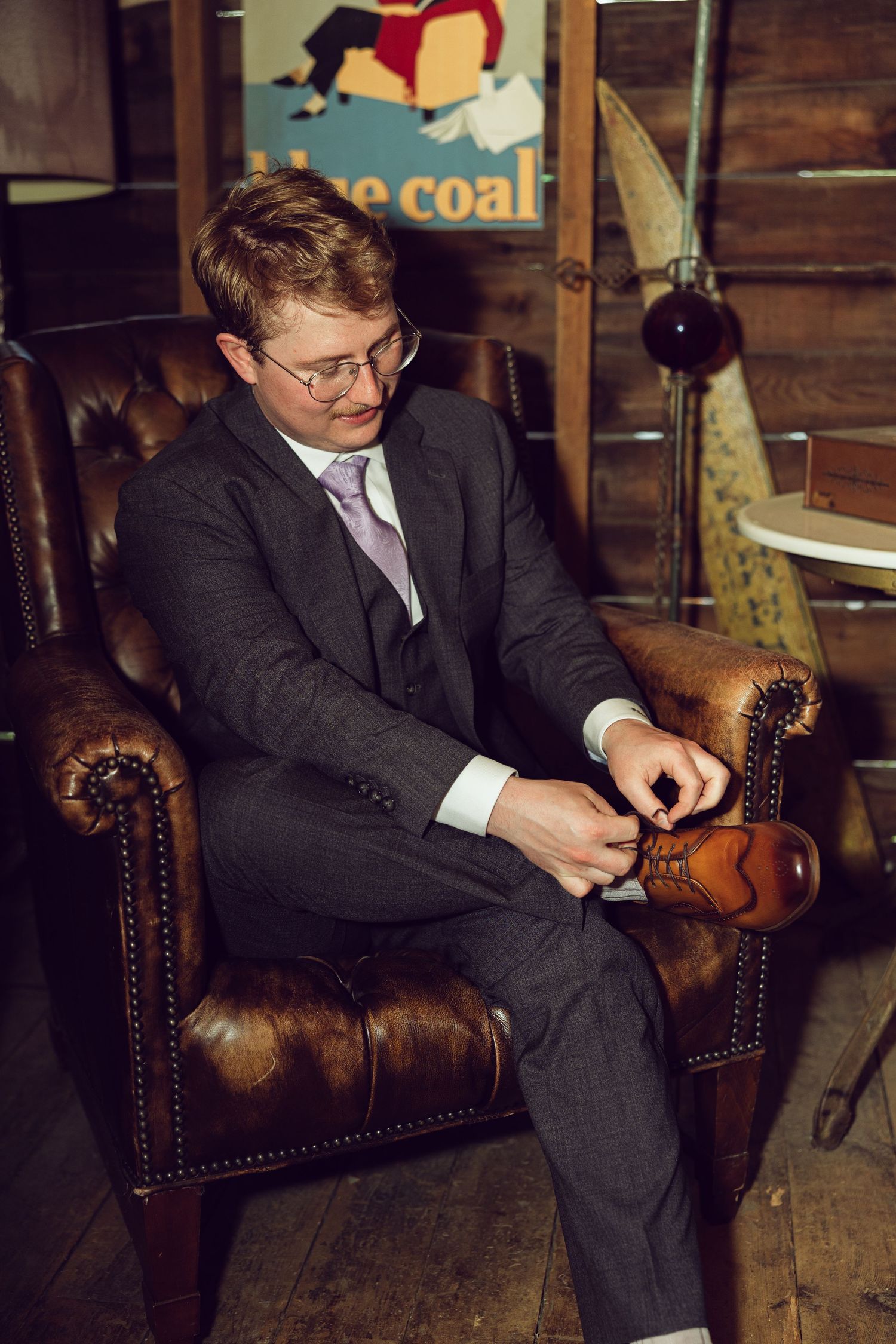 Groom in a gray suit and lavender tie sits in a tufted brown leather armchair, adjusting his tan dress shoe.