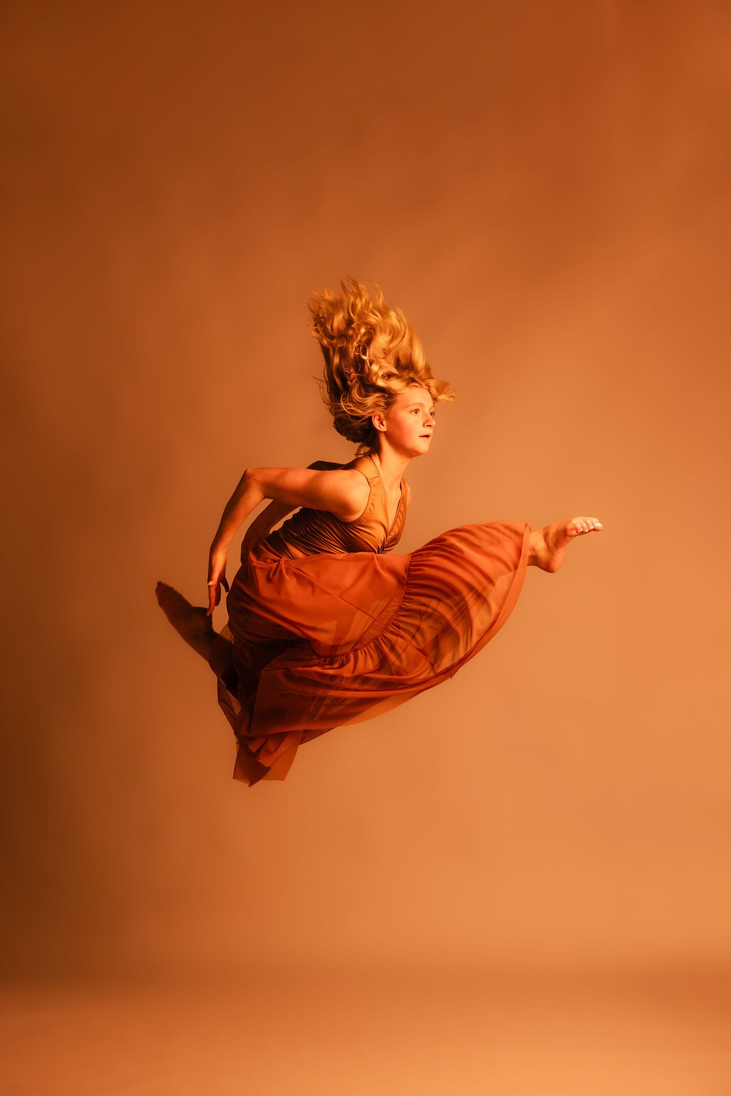 Dancer in a flowing orange skirt performs a mid-air split leap against a warm orange studio background.