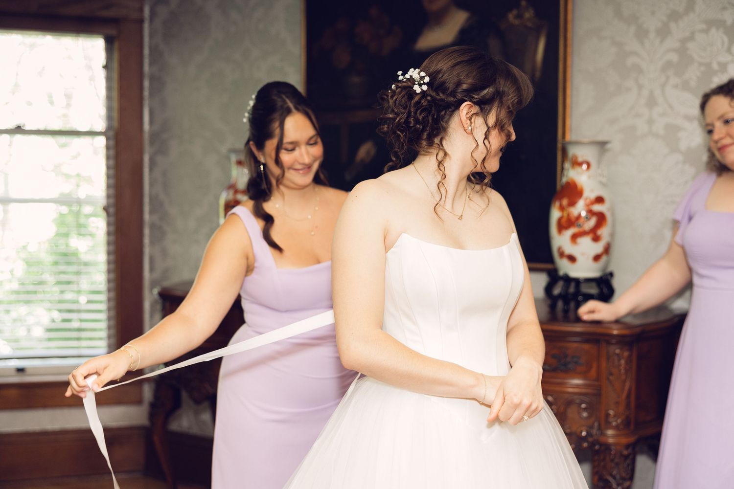 Bridesmaid in lavender helps tie the bride's white wedding dress in a vintage room with floral wallpaper and an ornate vase.