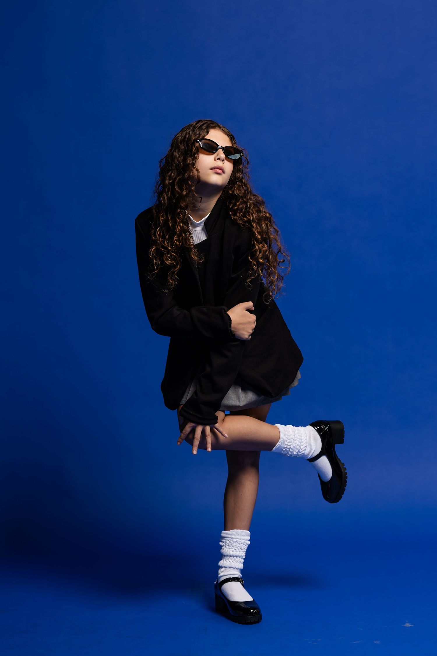 A girl with long curly hair, sunglasses, black blazer and white socks poses on one leg against a vivid blue backdrop.